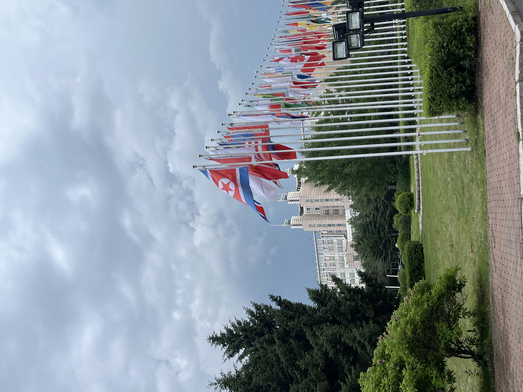 The image captures an outdoor scene featuring a row of national flags, predominantly the flag of North Korea at the forefront, followed by a long line of other flags on white poles. In the background, a large, multi-story building with a modern architectural style is visible, characterized by rows of windows and a light-colored facade. Adjacent to the building are evergreen trees and meticulously manicured bushes. The ground in the foreground is paved with brick. The sky is overcast with heavy, gray clouds. This scene is located at the Changchun World Sculpture Park in Changchun City, China, which is known for its collection of international sculptures and the display of various national flags.
