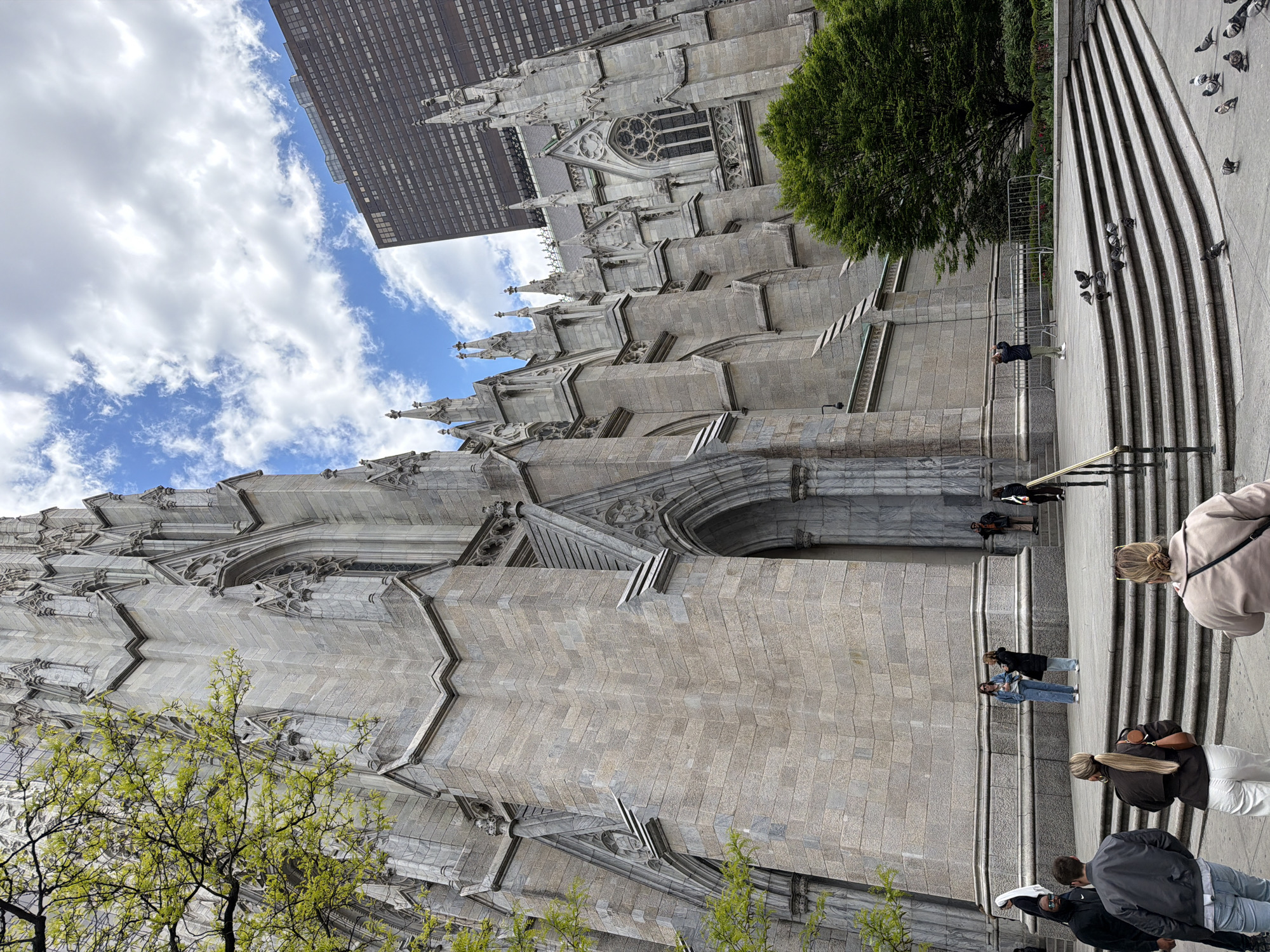 The image depicts St. Patrick's Cathedral in New York City. The architecture of the cathedral is visible in detail, featuring stone construction, arched entrances, and ornate spires. A modern skyscraper stands in the background, highlighting the contrast between historical and contemporary architecture. People are seen on the steps of the cathedral, some interacting with each other, while others are observing or photographing the building. Pigeons are present on the steps. The sky is partly cloudy. Vegetation, including trees, frames the cathedral, adding a natural element to the urban landscape.