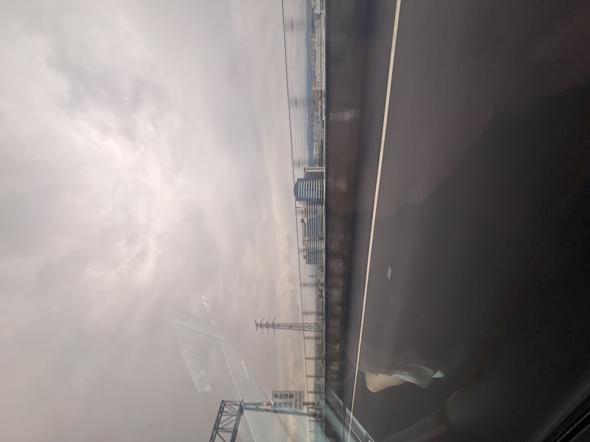 The image captures a view from inside a vehicle, likely a car or bus, looking out towards a cloudy sky and a cityscape. The foreground shows a wet, reflective asphalt road with a white line marking a lane. In the mid-ground, there is a barrier or railing separating the road from a lower level, possibly a bridge or elevated highway. Beyond this, a tall, latticed electrical pylon stands prominently. Further in the distance, a cluster of modern buildings with glass facades can be seen, indicative of a commercial or business district. The sky is overcast, with diffused light suggesting a cloudy or possibly foggy day. The overall impression is of an urban environment viewed during transit, with reflections on the glass creating a sense of depth and layering within the image. There is no clear indication of the specific location being Guangzhou City, China, based solely on the visual elements presented.