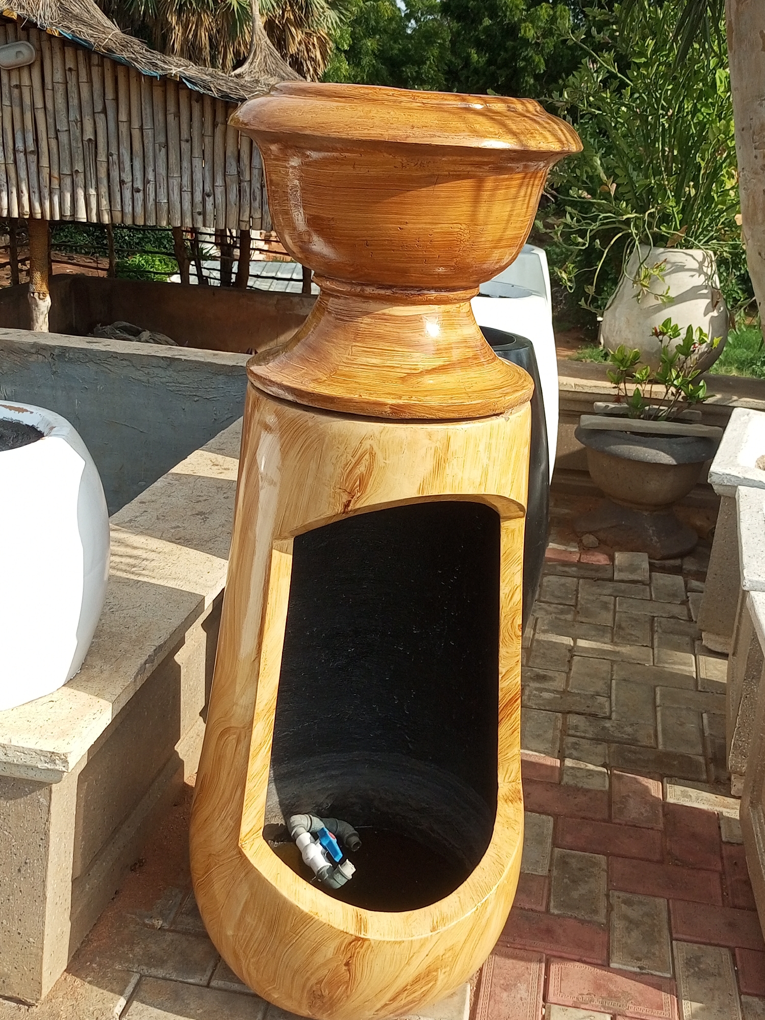 This media file depicts an outdoor scene with several decorative objects, likely in a garden or landscaping display area. The central focus is a large, ornate planter with a wood-grain finish, shaped like an urn atop a pedestal. The planter has a wide, flared opening at the top and a hollow, elongated base with an open cavity. Inside this cavity, a small section of plumbing, including a blue and white valve, is visible.
To the left of the central planter, a portion of a large, white, rounded planter is visible. Behind the planters, a thatched-roof structure, possibly a hut or gazebo made of bamboo, is present, suggesting a rustic or tropical setting. Further in the background, lush green foliage and trees are visible, along with other decorative pots. The ground is paved with interlocking brick pavers in shades of red and gray.
The lighting suggests it is daytime, with strong sunlight casting distinct shadows, indicating a bright and sunny day. The overall scene conveys a sense of outdoor decoration and potentially a display of garden accessories. No people are visible in the frame. There is no visible text in the image. The location context of Gombe, Nigeria, suggests a warm, possibly tropical climate.