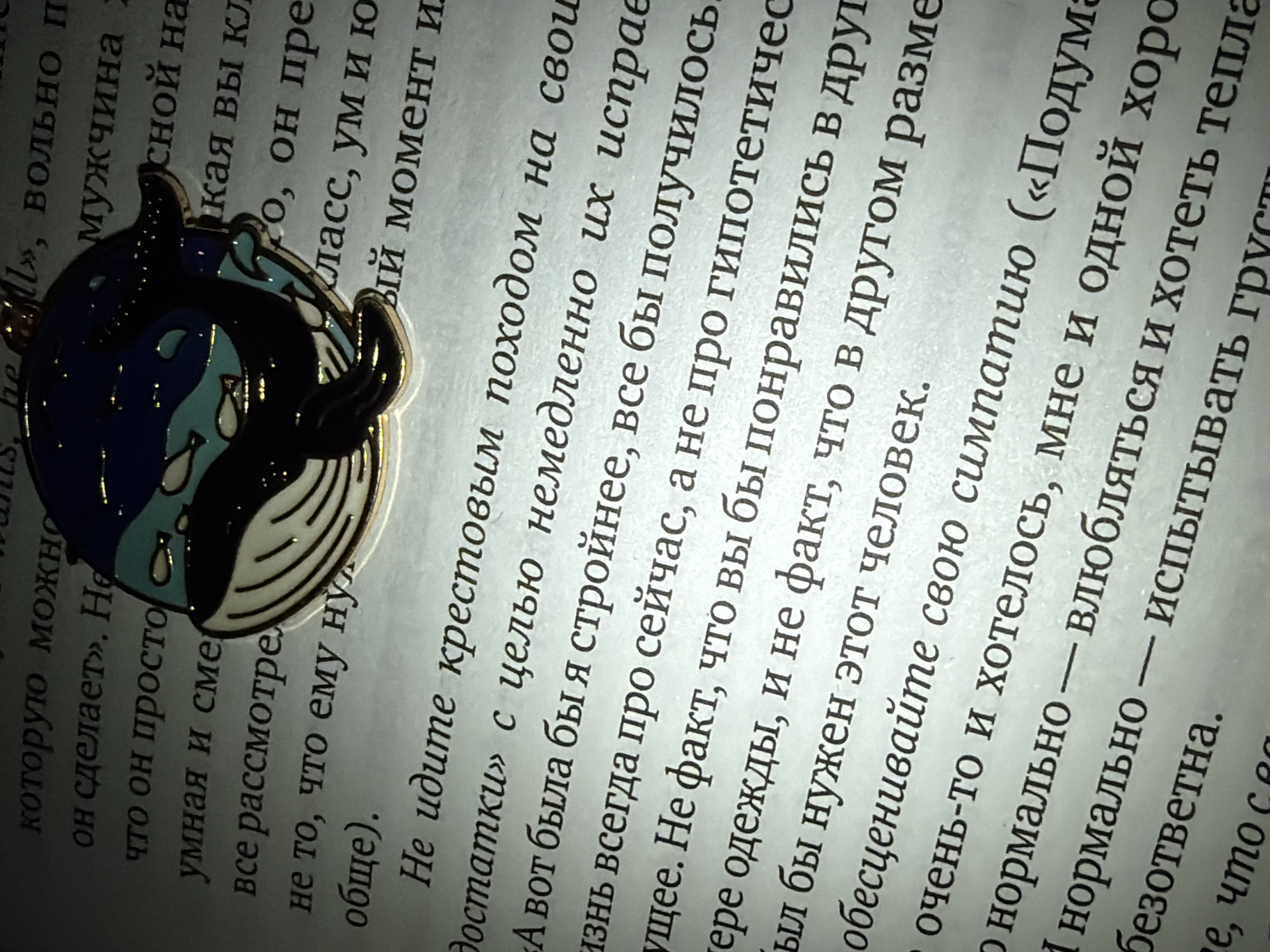 The image shows a book with Russian text and a whale-shaped pin on top of it. The pin is gold-colored with a black top half and a white bottom half. The black half has blue details resembling water, and there are teardrop shapes. The book's text is in focus near the pin, while the surrounding areas are blurry. The scene is indoors, likely on a table or another flat surface.