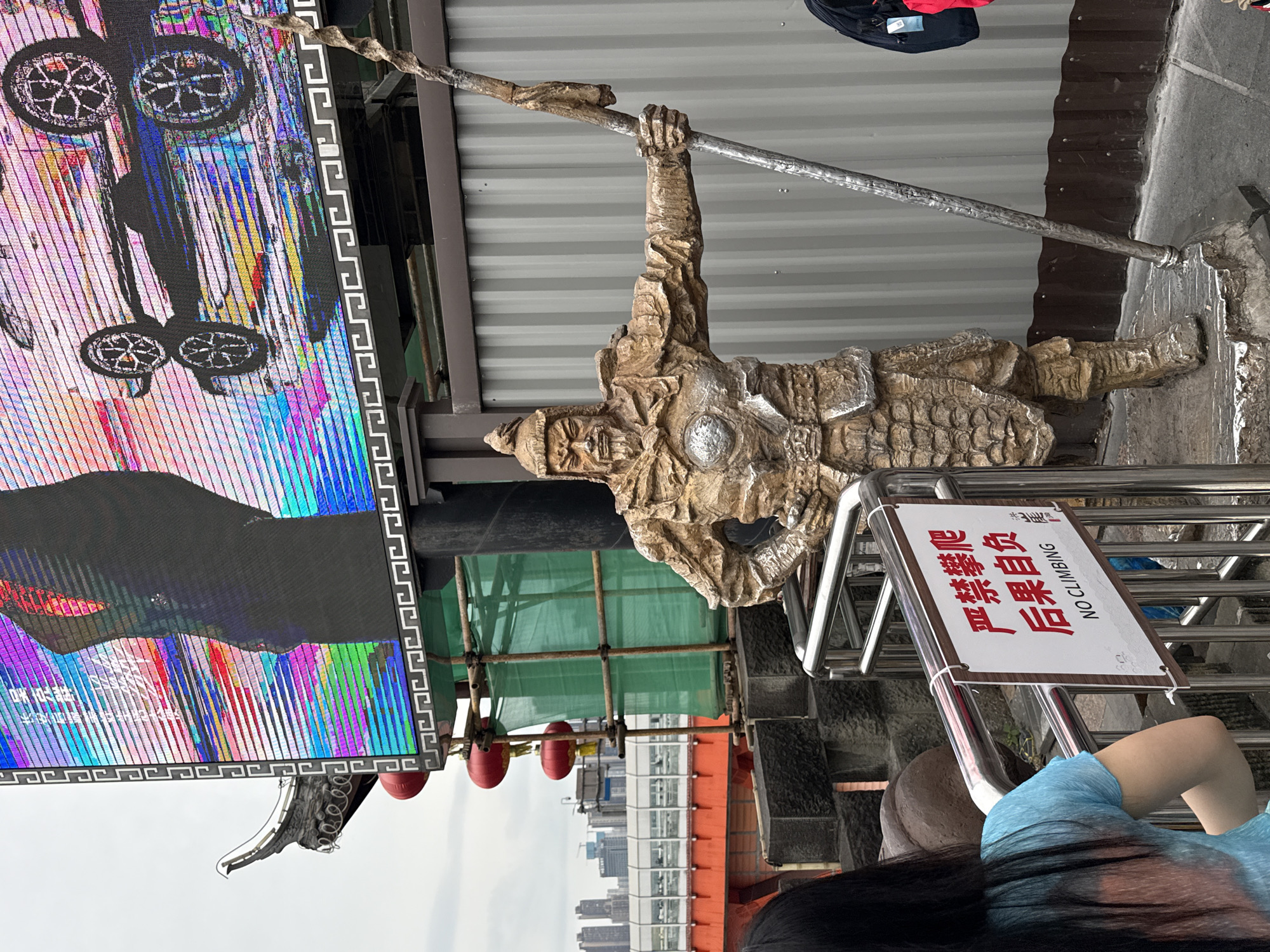 The image shows a sculpture of a warrior figure in Yuzhong District, China. The warrior is positioned hanging from a horizontal bar, possibly made of wood. A large LED screen is visible to the left, displaying an abstract image. A sign with the text "NO CLIMBING" is present, indicating that the sculpture is not intended for interaction. In the background, a cityscape is visible, suggesting an elevated location. A person wearing a blue shirt is partially visible in the foreground, and the edge of their long black hair.