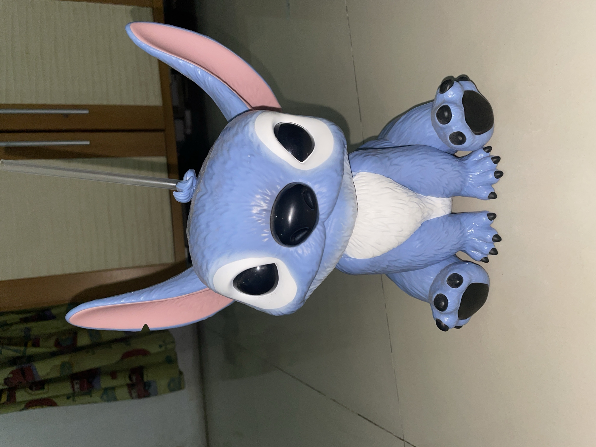 A plush toy resembling the character "Stitch" from the Disney film "Lilo & Stitch" is centrally located, oriented vertically on a tiled floor. The toy is blue with white fur on its chest and pink inner ears. It has large, black eyes and a large, black nose. A transparent plastic rod is attached to its back, extending toward the upper left corner. In the background, partially visible, are a wooden panel and a curtain with a printed pattern. The location is consistent with a domestic interior in North Jakarta, Indonesia, given the likely composition of materials and ambient light. No people are present.
