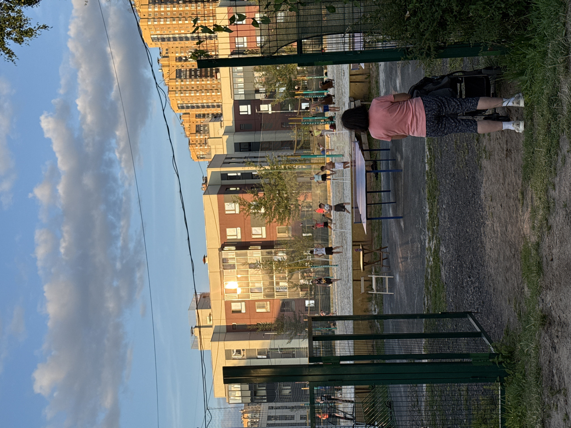The image depicts a sandy sports court with a volleyball net, located in a residential area of Pargolovo, Russia, at dusk. Several individuals, including adults and children, are engaged in recreational activities on the court. A woman stands in the foreground, facing away from the camera, pushing a stroller. Behind her, a tall, modern apartment building dominates the skyline, with a row of similar buildings stretching into the distance. The sky is partially cloudy, with the setting sun casting a warm glow on the scene. The court is surrounded by a fence, and a gate in the foreground provides access to the area. Tables and chairs are set up near the court, suggesting a communal gathering space. The overall atmosphere is one of leisure and community.
