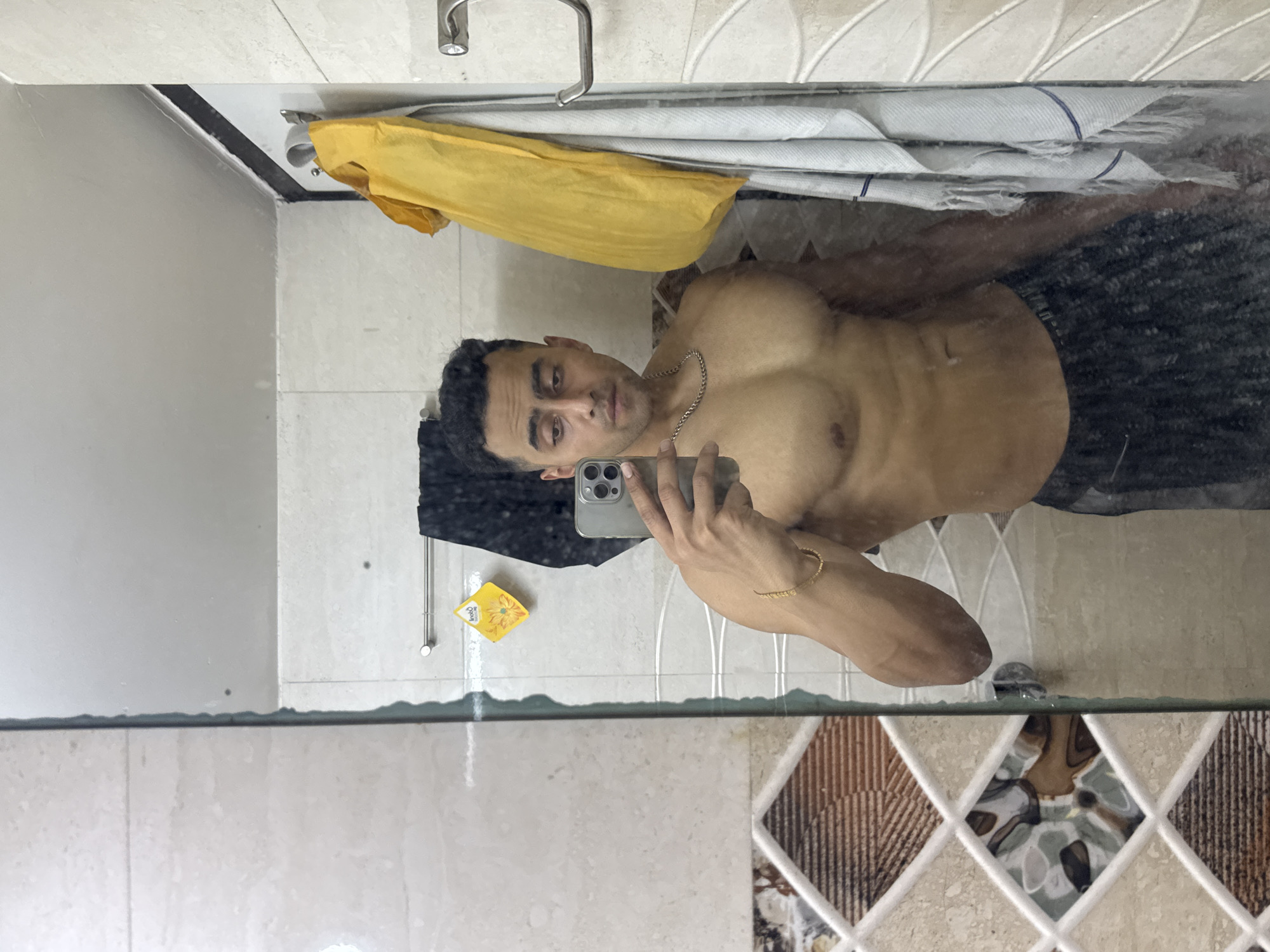 A man is taking a selfie in a bathroom. He is shirtless and wearing shorts. He is holding a smartphone in his left hand. He has a gold bracelet on his left wrist and a silver chain around his neck. The bathroom has white tiles and a sink. There is a yellow towel hanging above the sink. The location is Kolkata, India.