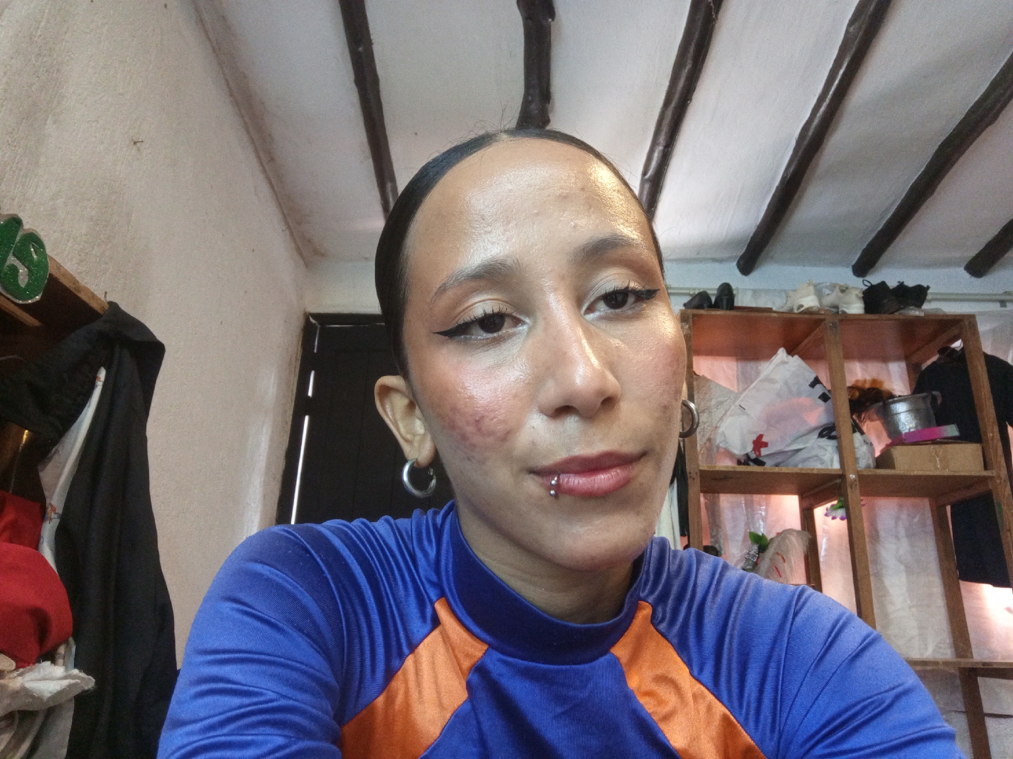 A person is facing the camera in a selfie pose. They have dark hair pulled back, dark eyeliner, and a lip piercing. They are wearing a blue and orange athletic shirt. Behind them is a room with white walls and a ceiling with exposed dark wooden beams. There's a wooden shelf unit with various items on it, and a garment rack with clothes hanging. The location cannot be determined solely from this image.