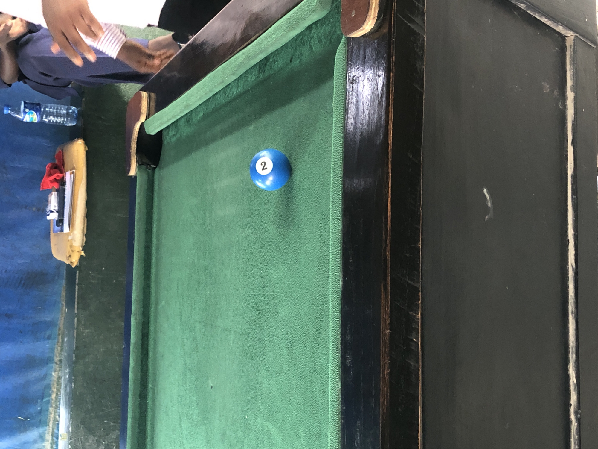 This image captures an indoor scene featuring a billiard table in the city of Unknown, Unknown. The central focus is a green-felted pool table, which exhibits signs of use and wear, including slight indentations and variations in the felt texture. A single blue billiard ball with the number '2' prominently displayed in white is positioned on the playing surface, slightly right of center. The table's rails and frame are constructed from dark wood, showing scuffs, scratches, and some minor paint chipping. A wooden bridge cue head rests on the far right rail of the table.
In the background, behind the pool table, there is a dark green raised ledge or platform. Above this platform, a blue wall with a rough texture, possibly concrete, shows discoloration and signs of age. On the left side of the green platform, a yellow-cushioned stool or bench holds a white notebook, a folded red cloth, and a small white object, possibly an electronic device. To the right of the stool, a clear plastic bottle containing blue liquid, likely water, stands on the green platform.
Two individuals are partially visible on the right side of the frame. One person, with dark skin, is seated in dark blue trousers, with their hands visible near their lap. The second person, also with dark skin, is standing closer to the camera, wearing a white shirt with thin horizontal stripes on the sleeve and dark trousers. Their right arm and hand are extended towards the table, suggesting observation or an impending action related to the game. The overall environment appears well-used and functional, consistent with a casual game room setting.