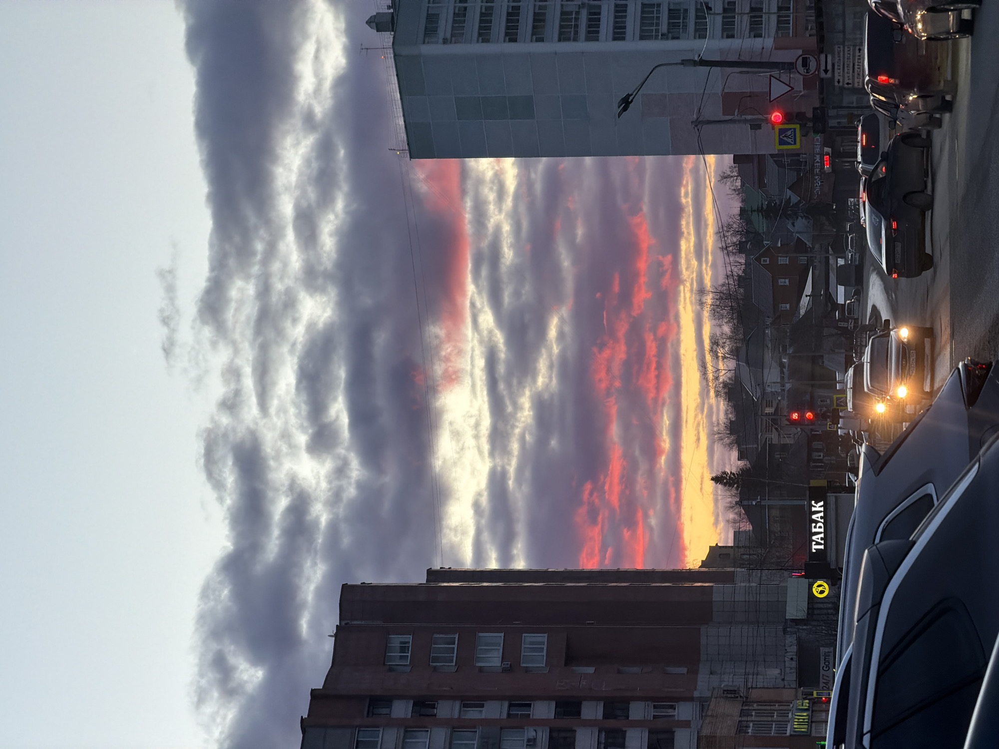 The image depicts a street scene in Perm, Russia at sunset. The sky is filled with clouds illuminated by red and orange hues, visible between tall buildings. A "TABAK" store sign is visible on the right. Cars are stopped at a traffic light, showing red. A pedestrian crossing sign is visible. A tall building is on the left, another brick building underneath it.