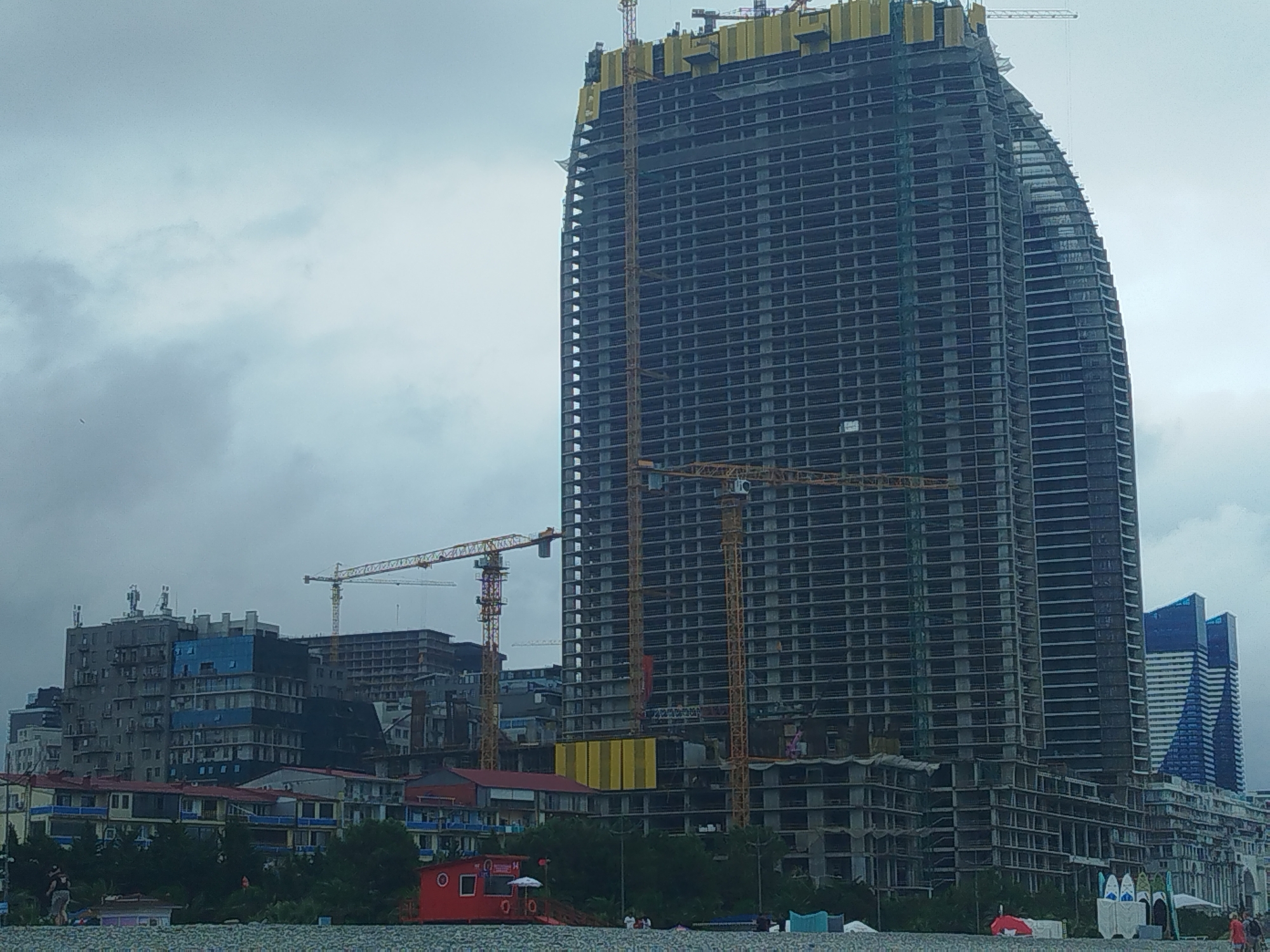 The image shows a construction site in Batumi, Georgia, with several tall buildings under construction. A prominent, unfinished skyscraper with a dark facade and visible floors dominates the center of the frame. Multiple yellow construction cranes are positioned around the skyscraper, indicating ongoing work. To the left of the skyscraper, other buildings, some appearing to be residential or hotels, are visible, with varying levels of completion and architectural styles. In the foreground, a strip of land with trees and some low-lying structures, possibly part of a beachfront, is present. The sky is overcast with clouds. There are a few indistinct figures of people present on the beach area. The scene captures a developing urban landscape with a focus on large-scale construction projects.