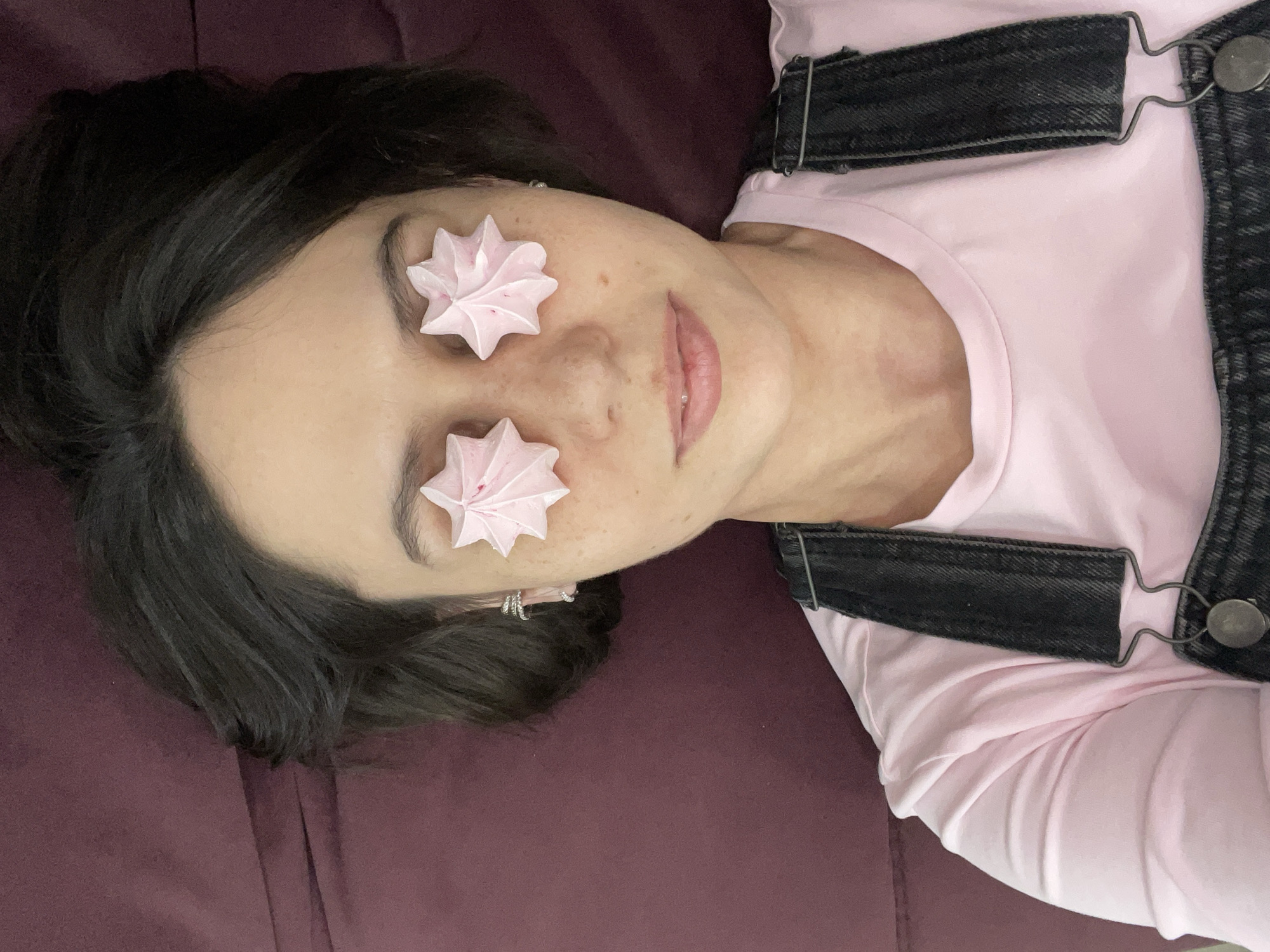 A young person with dark hair is depicted lying on a dark reddish-purple surface, possibly a bed or sofa. Their head is positioned towards the upper left of the frame, with their body extending downwards to the right. They are wearing a light pink t-shirt underneath dark denim overalls, visible from the shoulders and chest area.
Two light pink, star-shaped meringue cookies are placed on the person's face, covering their closed eyelids. Their mouth is slightly open, revealing light pink lips. A small silver hoop earring is visible in their right earlobe (viewer's left). The overall pose and use of meringues suggest a playful or whimsical moment of rest or relaxation. The lighting appears soft and even, indicating an indoor setting. There is no visible text in the image. This scene is contextualized as being from Izhevsk, Russia.