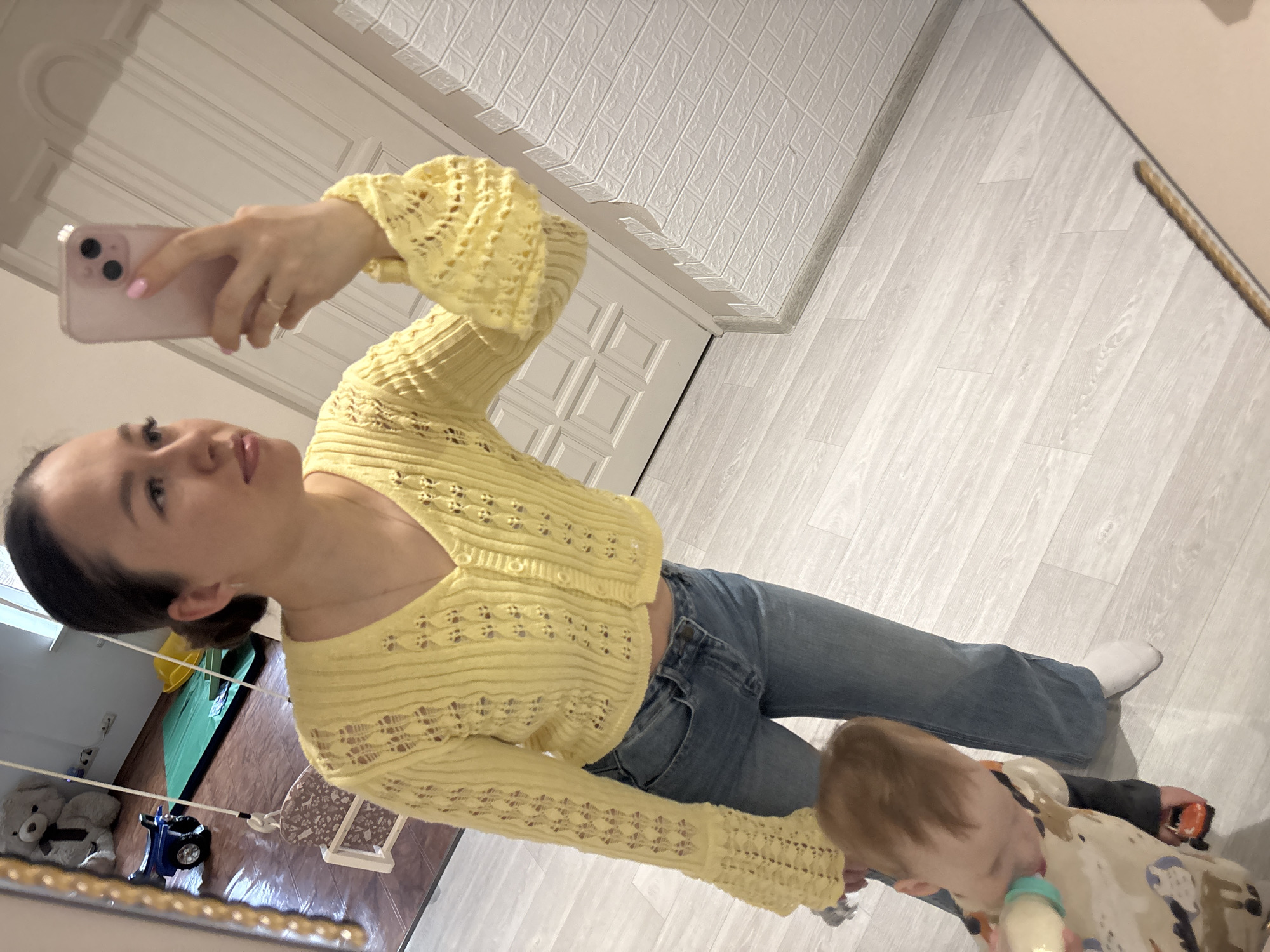 A full-length mirror selfie captures a woman and a child in what appears to be a home interior in Ферма, Russia.
The primary subject is a woman, seen from the waist up in the mirror's reflection, holding a light pink smartphone in her right hand to take the photo. She has dark hair pulled back and is wearing a light yellow, open-knit cardigan over what appears to be a light-colored top, paired with blue jeans. Her nails are painted a soft pink. She looks into the camera with a neutral expression.
Standing next to her, partially visible, is a young child with light brown hair, wearing a patterned garment. The child is focused on drinking from a baby bottle, held in both hands.
The room features light-colored, wood-laminate flooring and a ceiling with white, textured tiles that resemble bricks. A white door with raised panels is visible in the background. Other elements glimpsed in the reflection include a stuffed teddy bear, a small blue toy vehicle, and parts of a dark-colored surface (possibly another piece of furniture or an area with a different flooring type) which reflects more light. The overall lighting suggests it is daytime. No text is visible in the image. The scene captures a casual, everyday moment within a home.