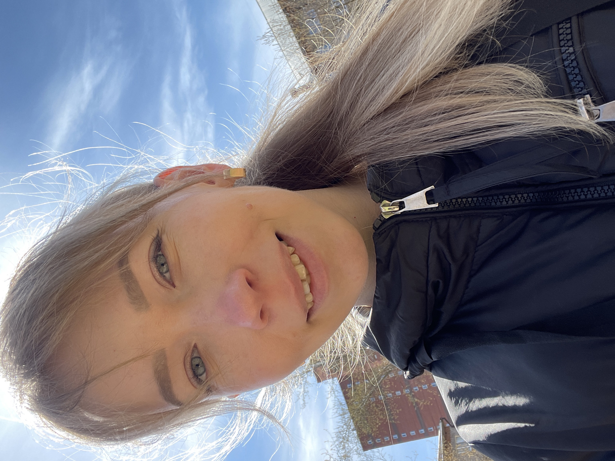 A young woman with light blonde hair and blue-green eyes is taking a selfie outdoors. She is wearing a black puffy jacket with a silver zipper. The background shows a bright blue sky with some white clouds. A glimpse of a brick building and some trees can be seen, suggesting an urban setting. Given the information about Perm, Russia, it can be inferred that the photo was taken in that city. The sun is shining brightly, illuminating the woman's face. She is smiling slightly. A gold-colored ring is visible on her ear.