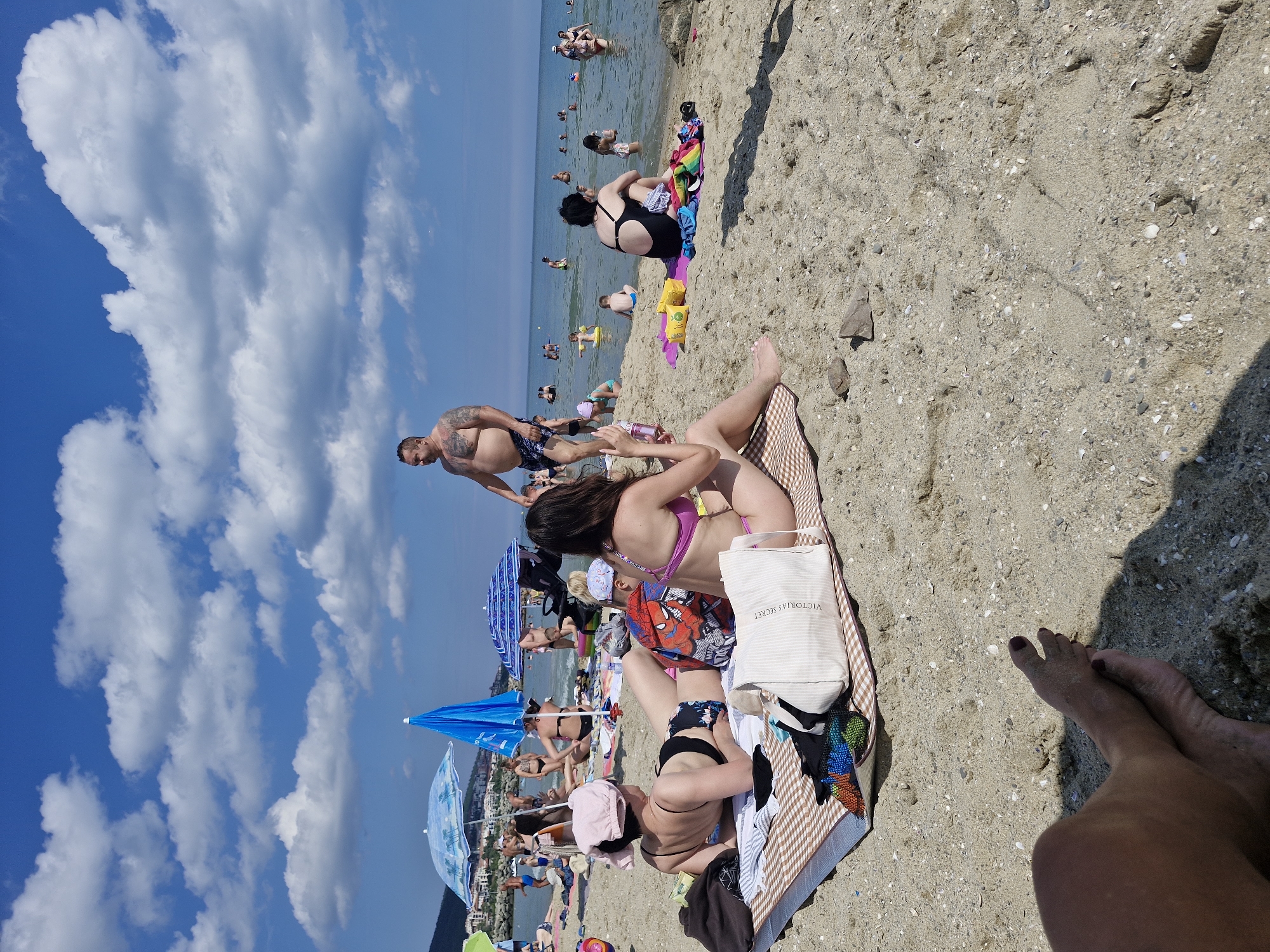 A sunny day at Sveti Vlas, Bulgaria, unfolds on a sandy beach with numerous people enjoying the sea and sun. In the foreground, a person's legs and feet, with dark nail polish, are resting on the sand, casting a shadow. Further back, two women are seated on a checkered blanket. One, wearing a pink cap and black bikini bottoms with patterned fabric, is lying down, facing away from the camera. Beside her, a woman in a pink bikini is sitting, also facing away. A child's backpack with a Spider-Man design is placed between them. A white tote bag with "VICTORIA'S SECRET" visible rests on the blanket.
In the middle ground, a man with tattoos on his chest and arms, wearing dark patterned swim trunks, stands and appears to be speaking with someone off-camera. Behind him, several beach umbrellas, one with a blue and white palm tree design and another solid blue, are erected. Many people are scattered across the beach and in the shallow water. Some are standing, some are sitting, and children are playing. The sea is calm, with people swimming and wading. In the background, buildings and hills, characteristic of Sveti Vlas, are visible along the coastline. The sky is a vibrant blue, dotted with fluffy white clouds.