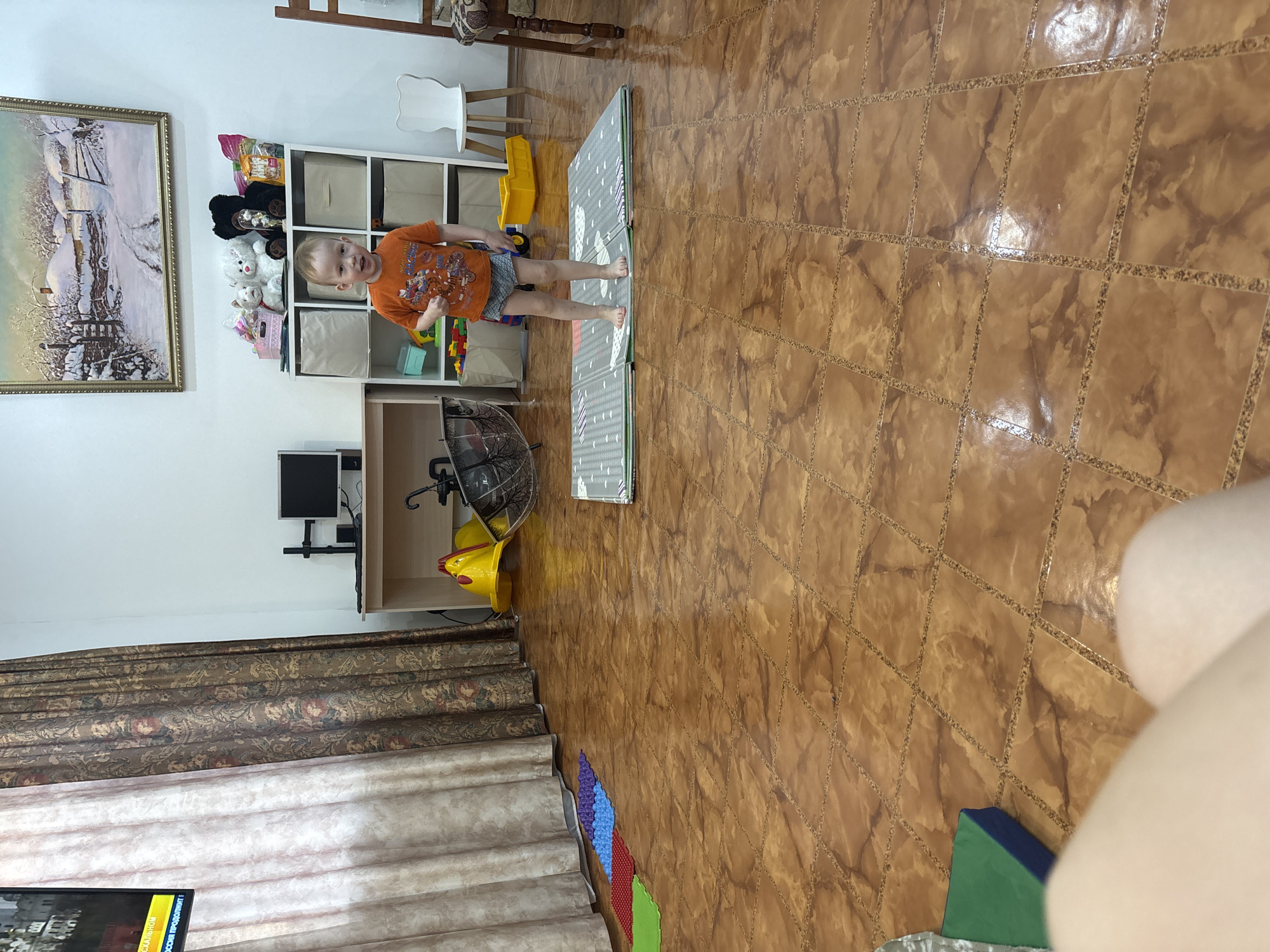 Here is a description of the image:
The image is a medium shot taken indoors. A young child is standing on a gray and white play mat in a room with a brown tile floor. The child is fair-skinned, with blonde hair, and is wearing an orange t-shirt with a graphic design, and blue shorts.
Behind the child, there is a white shelving unit filled with toys and storage boxes. To the left, there is a framed painting on the wall. A desk with a small monitor is also visible in the background. There is a yellow toy duck on the floor near the desk.
The room has patterned curtains on one side. A portion of a TV screen is visible at the bottom left. The floor is brown tile with a grid-like pattern. In the foreground, there are blocks of green and blue near a white area.
Based on the metadata (location is Ферма, Russia).
