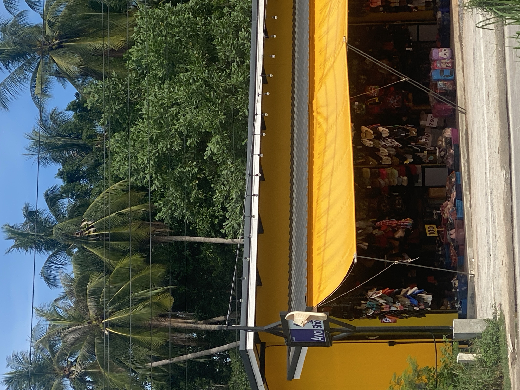 This outdoor shot depicts the exterior of a shop, possibly a souvenir store, located in Baan Nai Suan, Thailand. The shop is characterized by its bright yellow facade and a large, yellow awning. The store is filled with various merchandise, with prices indicated on some items, displayed on shelves. A sign with "Ava Sho" is visible. The scene is set against a backdrop of lush greenery, including palm trees and other trees. The road runs alongside the shop.
