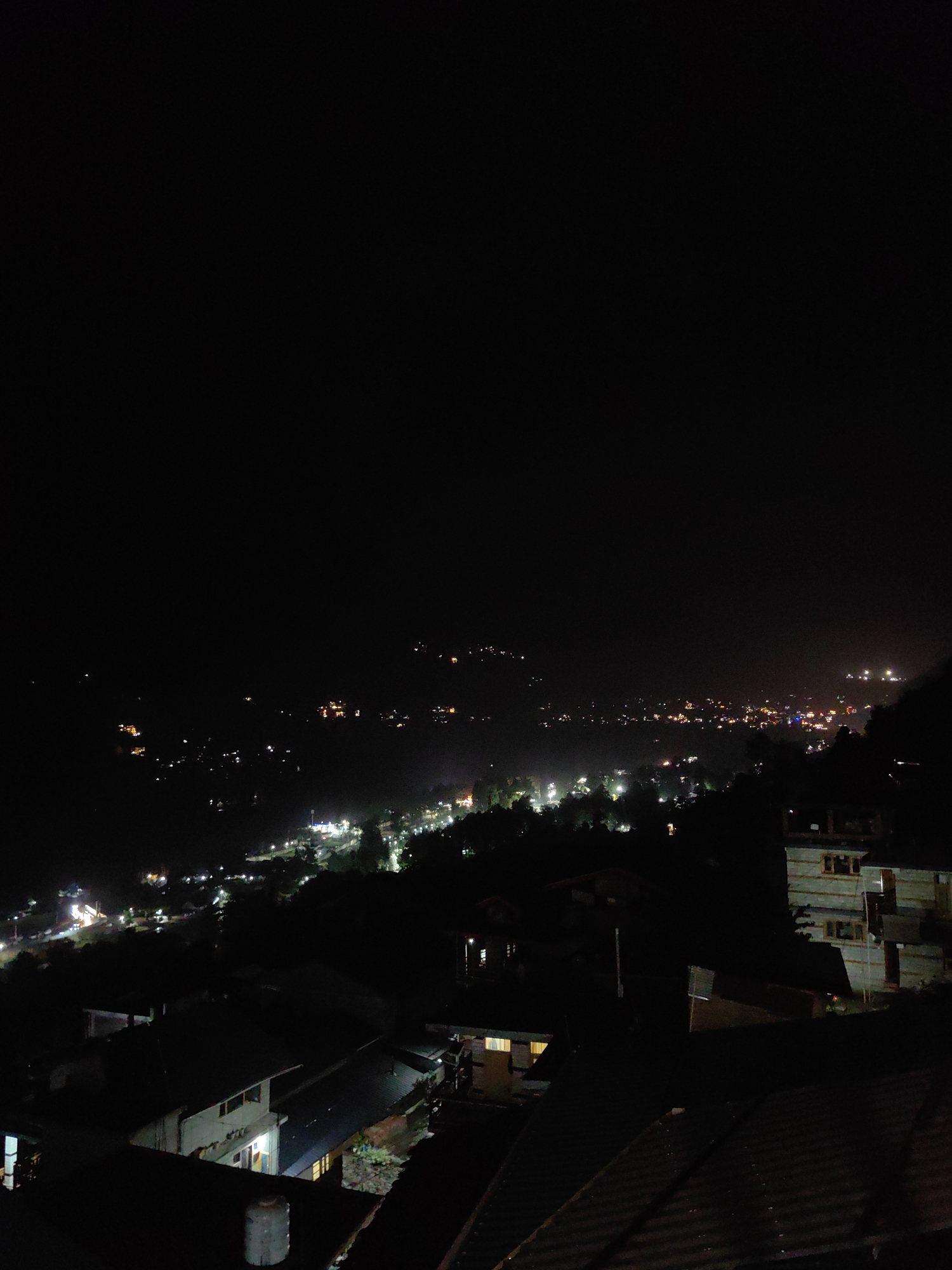 The image depicts a nighttime aerial view of Vashisht, India, a town known for its hot springs. The scene is characterized by darkness punctuated by numerous scattered lights from buildings and streetlamps, indicating a populated area nestled within a hilly or mountainous terrain. In the foreground, several rooftops and structures are visible, their details obscured by the low light. A water tank is present on one of the rooftops. Further into the midground and background, the lights become more dense, suggesting the core of the town. The overall impression is that of a remote settlement illuminated against the vast darkness of the night.