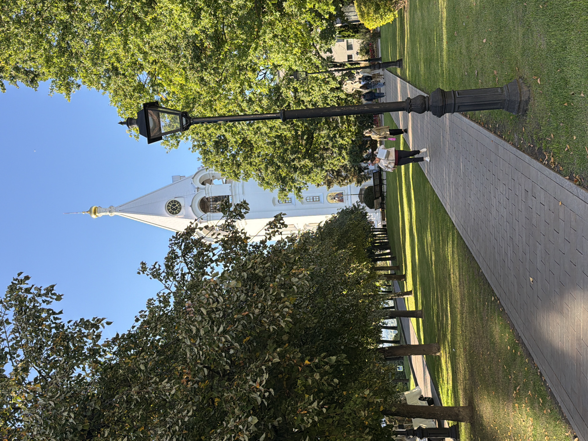 The image shows a view of the Nizhny Novgorod Kremlin in Russia. In the foreground, a paved pathway runs through a grassy park, lined with trees casting dappled shadows. Several people are walking along the path. A prominent white bell tower with a gold dome and a clock face rises above the trees in the background, along with other buildings of the Kremlin complex. A black lamppost stands beside the pathway in the foreground. The sky is clear and blue.