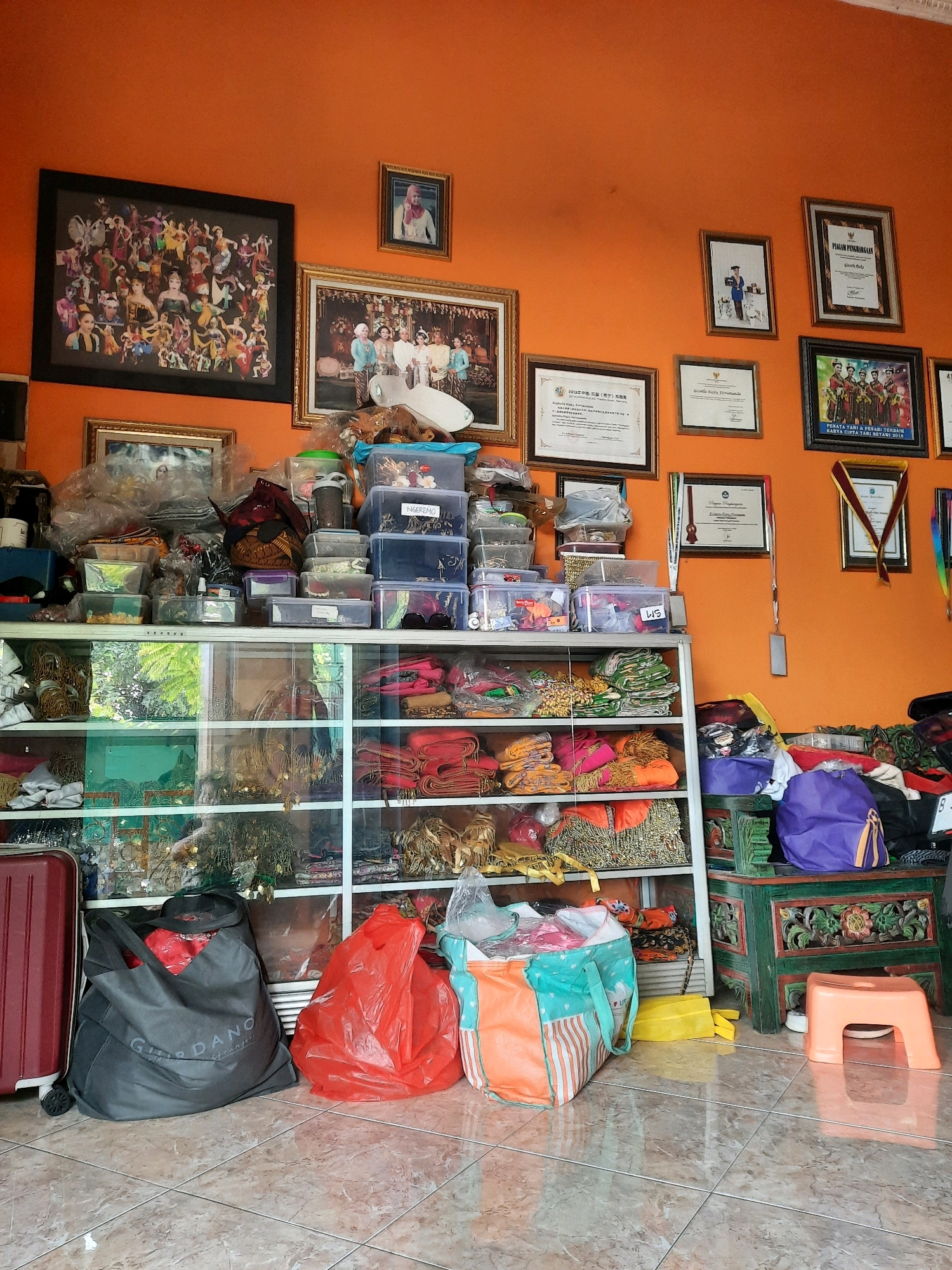 This is a medium shot, eye-level, indoor photograph taken in a room with orange walls. The room appears to be a shop or a storage area, filled with various items. In the center of the image, there is a white glass display cabinet with multiple shelves. These shelves are packed with folded fabrics, mostly in bright colors like red, orange, yellow, and green. There are also several clear plastic containers stacked on top of the cabinet, filled with smaller items and materials.
On the wall behind the cabinet, there are several framed pictures and certificates. The largest framed picture on the left depicts a group of people in traditional costumes. Other frames contain what appear to be certificates of achievement, with Indonesian text visible on some.
In the foreground, on the tiled floor, there are several bags. A large dark gray "Giordano" branded bag is on the left, partially filled with red items. To its right, a bright red plastic bag is crumpled on the floor. Another colorful tote bag with orange and teal stripes sits next to the red bag. A small orange plastic stool is also visible on the right side of the frame.
The overall impression is one of a well-stocked retail or storage space, possibly related to traditional Indonesian clothing or crafts, given the textiles and cultural imagery. The room's decor, particularly the orange walls and the style of the furniture and decorations, suggests a location within Indonesia, possibly in a residential or commercial area of East Jakarta.