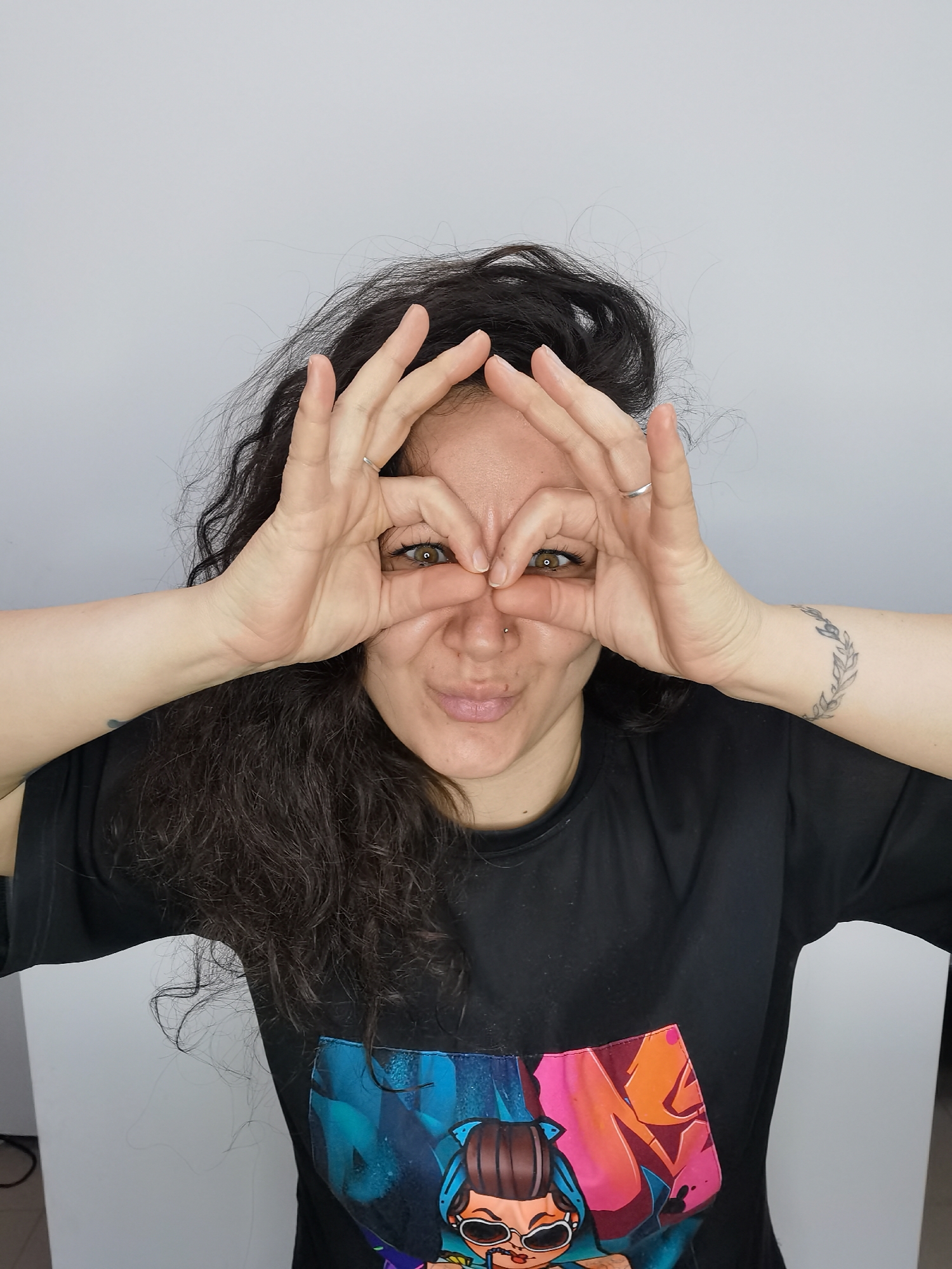 A fair-skinned woman with long, dark, curly hair is facing the camera, making eye-shaped circles with her hands around her eyes. She has a ring on the ring finger of her right hand, a nose piercing, and a tattoo on her left wrist. She is wearing a black t-shirt with a colorful graphic design on the front. The background is a plain, light gray or white wall. The location, Chaikovsky, Russia, is not evident from the image.