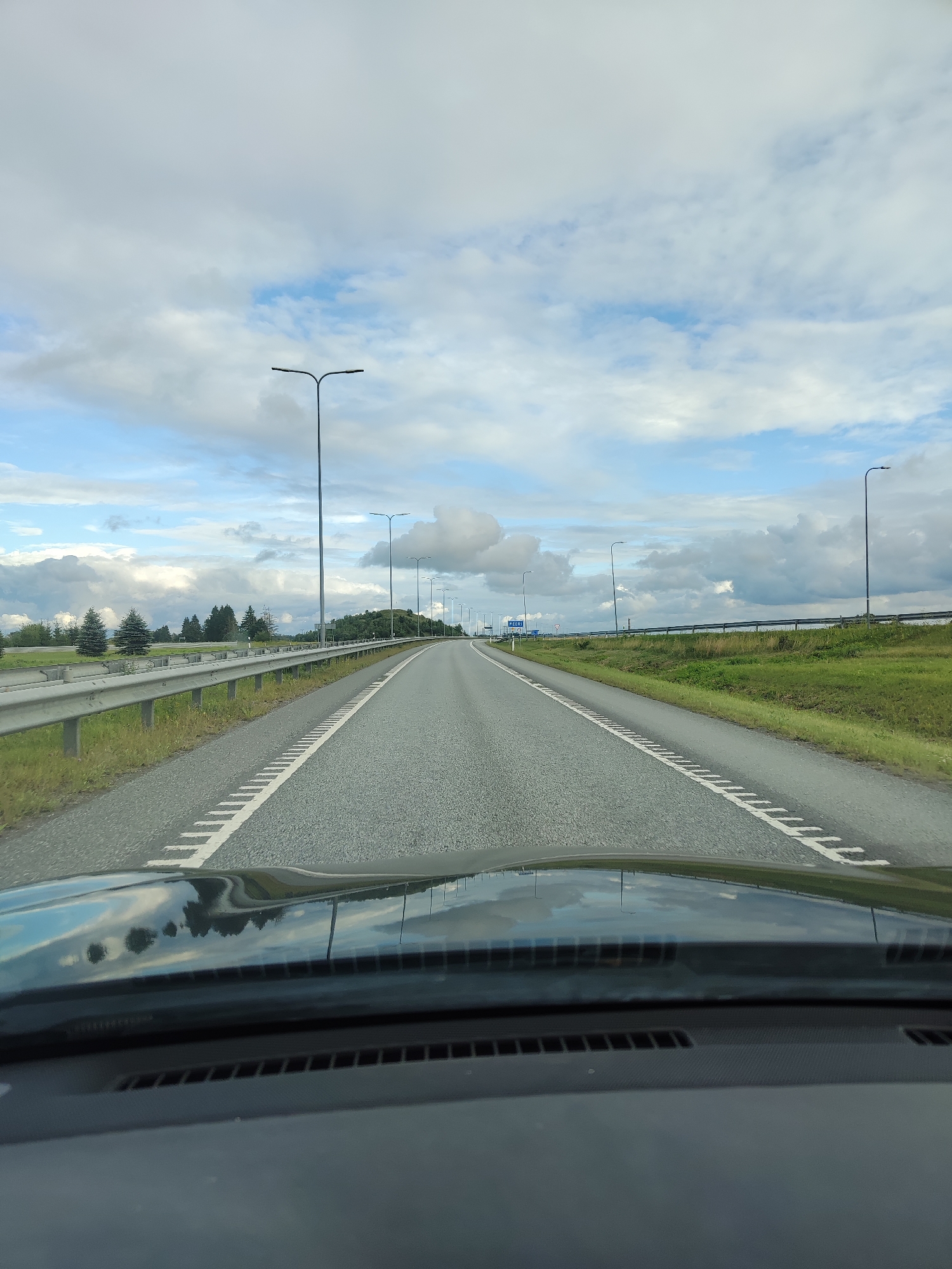 The image depicts a view from inside a vehicle traveling on a multi-lane highway. The highway stretches into the distance, flanked by grassy shoulders and a metal guardrail on the left. Several tall streetlights line the side of the road. The sky is partly cloudy with patches of blue. In the background, a low, tree-covered hill is visible on the right. A sign with blue text is visible on the left side of the highway in the distance. The dashboard of the car is in the foreground, reflecting the sky and trees. The scene appears to be in a rural or semi-rural area outside of a city. Based on the road markings and landscape, it is likely a highway in Estonia. The city of Kukruse, Estonia is a small settlement known for its industrial heritage and shale oil production, and while this image does not directly show any specific landmarks of Kukruse, it is plausible this highway is in its vicinity or leads to it.