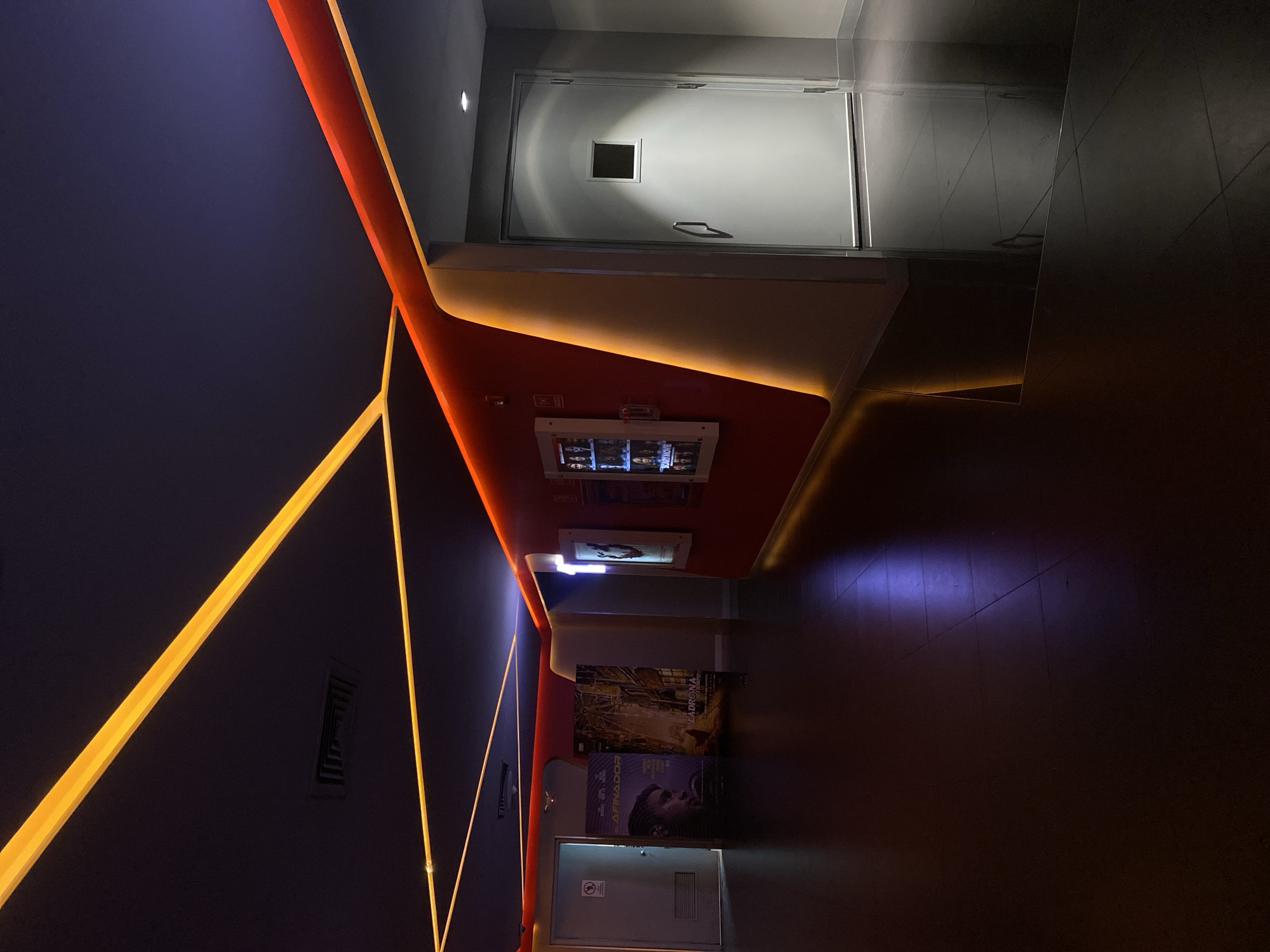 The image depicts an interior space, possibly a hallway in an entertainment venue, such as a movie theater, in Unknown, Unknown. The walls are dark, with geometric patterns created by bright yellow light strips. One wall features movie posters and promotional displays. A white door is visible at the end of the hallway, suggesting an exit or service area. The floor is dark and reflective. The overall lighting is dim, with the light strips and displays providing the main sources of illumination.