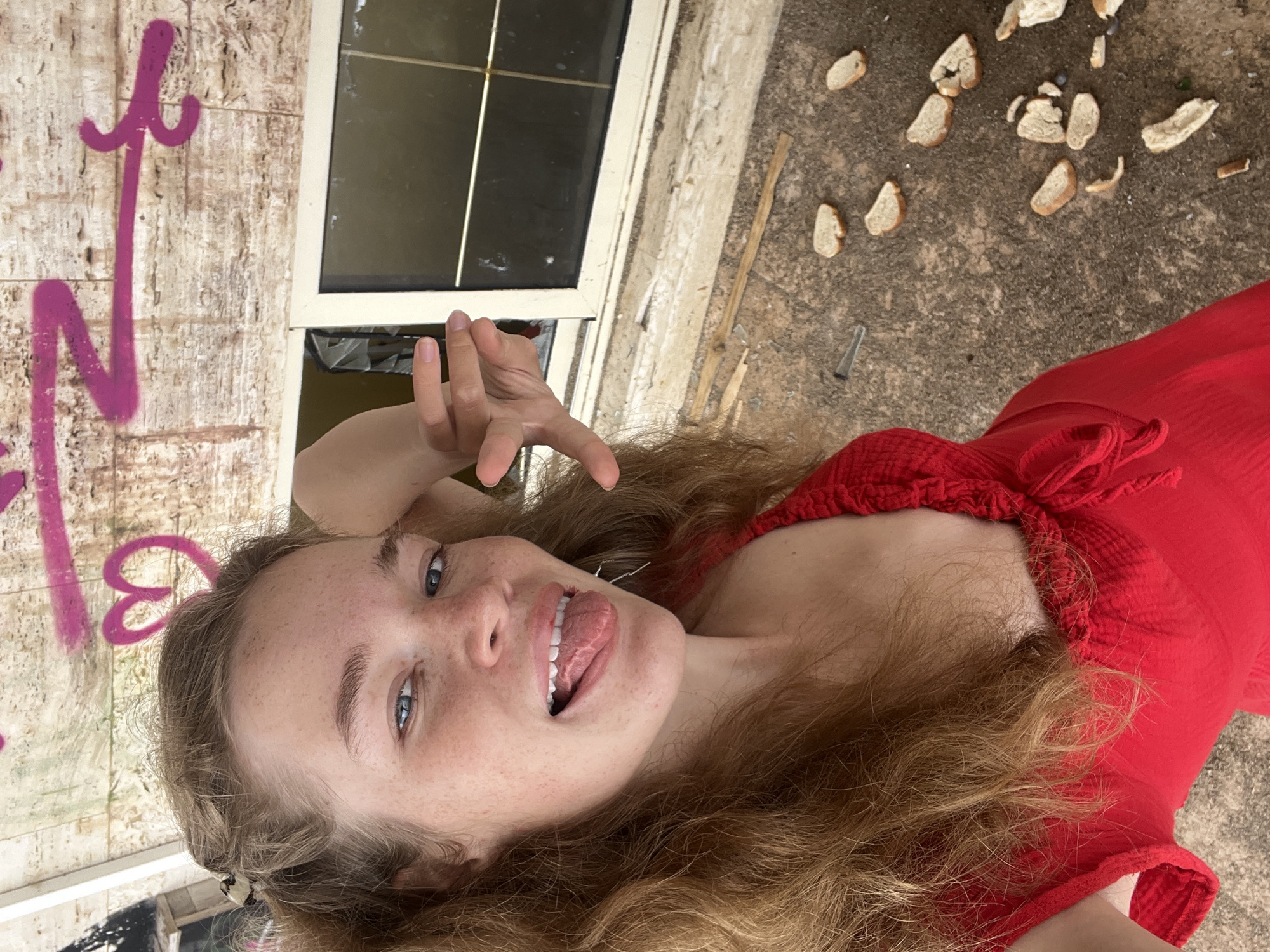 A young woman with long curly brown hair and freckled skin, wearing a red dress, sticks her tongue out at the camera and makes a "peace" sign. She is in an area with graffiti and a window and the ground nearby is littered with bread. The photo appears to be taken in Budva, Montenegro.