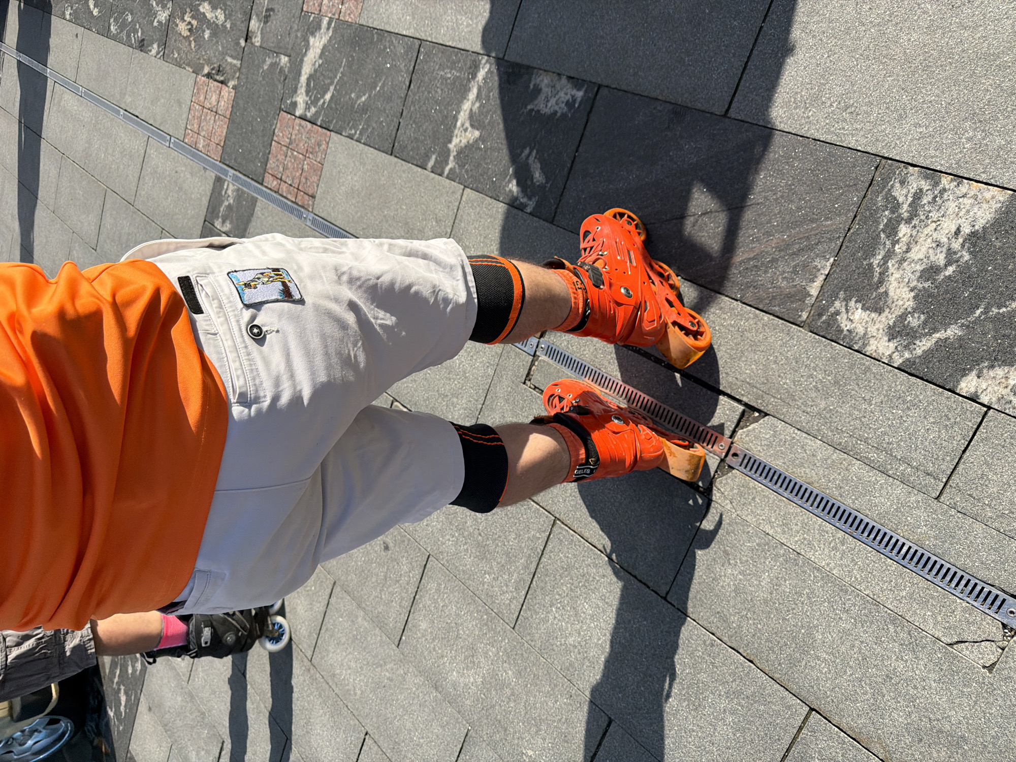 The image shows a person standing on a paved area, likely a sidewalk or plaza. The person is wearing an orange t-shirt, light-colored shorts with a patch, black and orange knee-high socks, and bright orange inline skates. The ground is paved with gray rectangular stones, and a metal grate drainage system runs along the paving. Shadows are cast on the ground. In the lower left corner, a second pair of rollerblades are partially visible. The location appears to be an urban area in Moscow, Russia.
