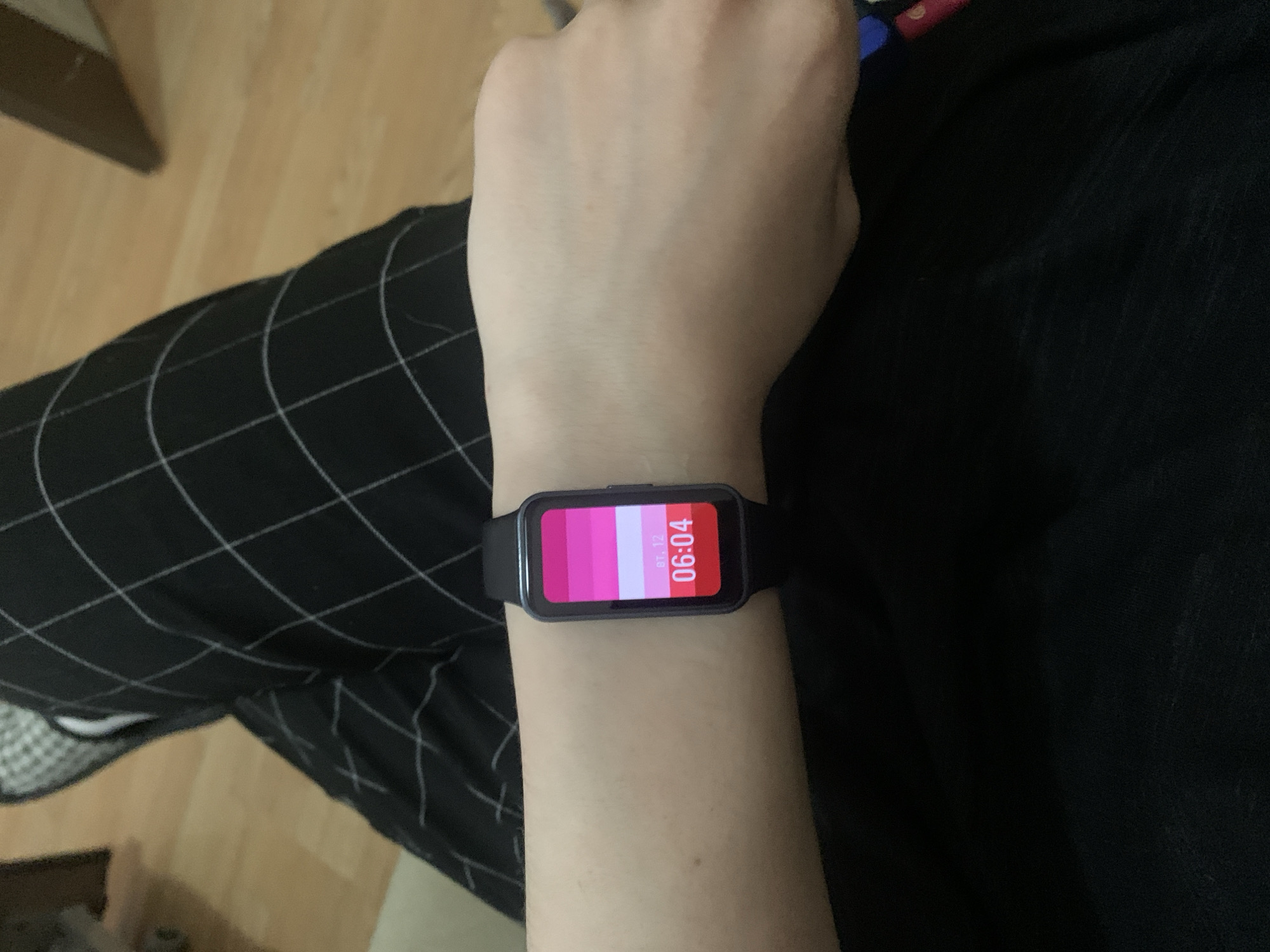 A person's left wrist, adorned with a black smart watch displaying a time of 06:04, is positioned in the center of the frame. The watch face exhibits a color gradient of pink shades. The wrist is situated against a backdrop of black clothing and black-and-white checkered pants. The environment includes a wooden floor. The location within the city of Unknown, Unknown is not discernible.