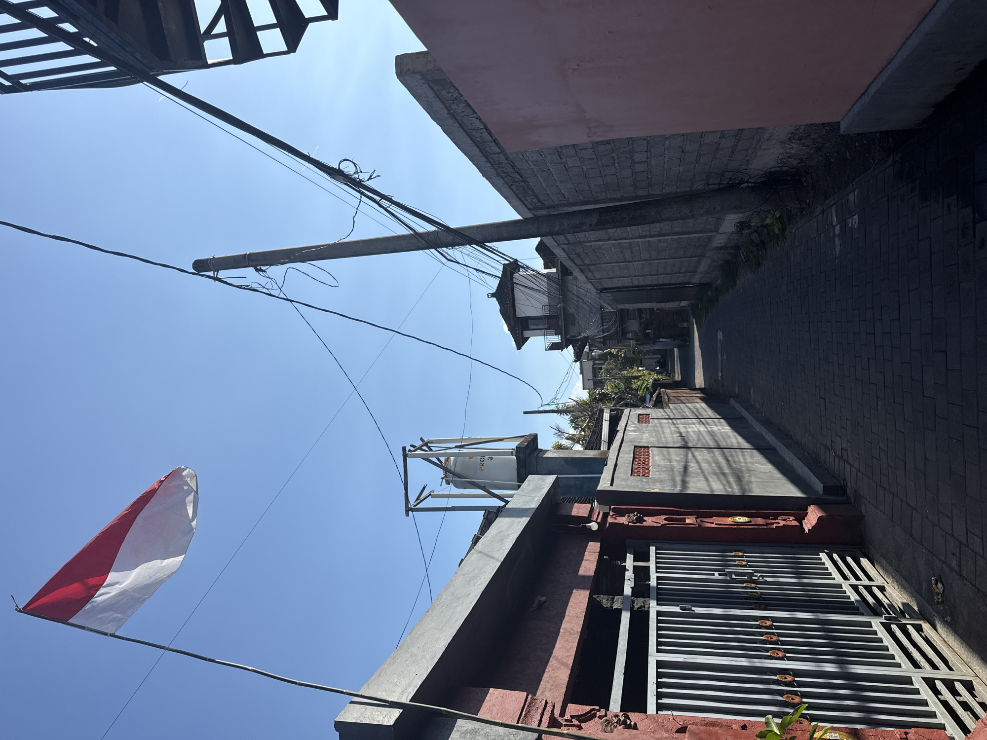 The image depicts a narrow alleyway in Denpasar, Indonesia, under a clear blue sky. An Indonesian flag is prominently displayed, attached to a bamboo pole, and unfurled against the sky. The alley is paved with dark rectangular bricks and is bordered by buildings and walls on either side. On the left, a building with a red facade features a metal gate with vertical slats and decorative circular elements. A water tank, labeled "PRO," is visible on top of this building. The right side of the alley is dominated by a grey brick wall and a pink-painted wall further down. Electrical wires crisscross above the alley, supported by a utility pole. In the distance, more buildings and a spiral staircase are visible. The scene appears to be a residential or commercial area, with no people present.