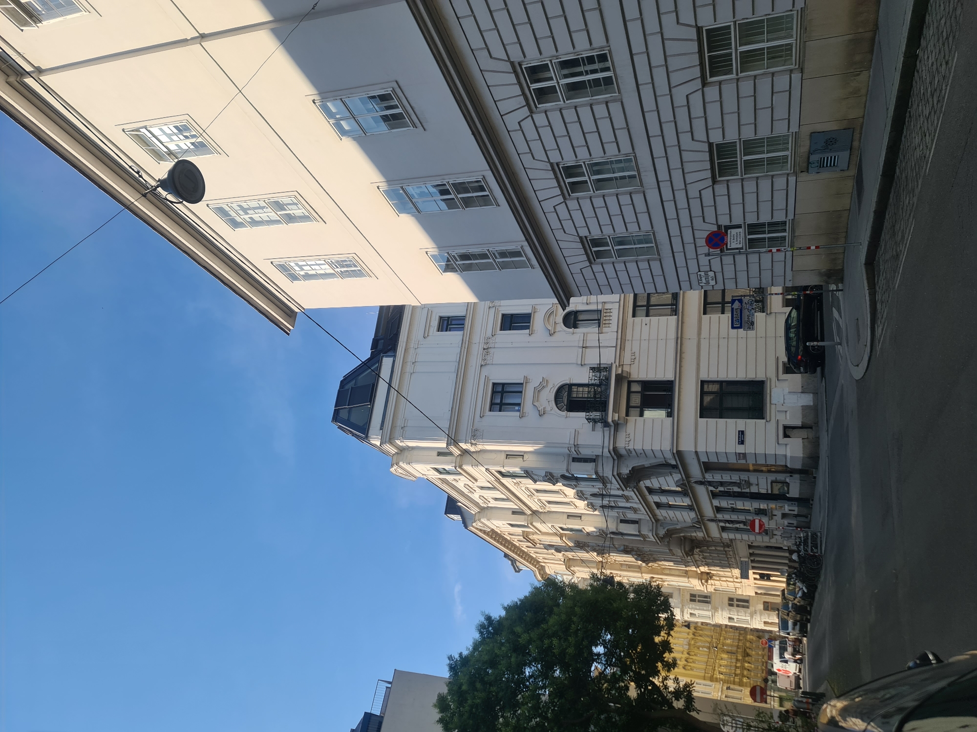 A street-level, eye-level, wide-angle photograph captures a view looking down a narrow street lined with white and cream-colored buildings with a clear blue sky above. The architecture suggests a European city, likely Vienna, Austria, with ornate facades, tall windows, and decorative elements. A prominent building on the right exhibits a light cream color with a shadow cast from the setting sun, highlighting its architectural details, including patterned stonework on the lower floors and numerous tall, multi-paned windows. A street lamp hangs from wires suspended across the street in front of this building.
To the left, a row of white buildings follows the curve of the street. One building towards the center has a distinctive dark, angled roof structure. Further down the street, older buildings with yellowish facades are visible. The street itself is paved, with a sidewalk on the right. Several parked cars, including a white van and a black sedan, are present. A bicycle rack is visible near the sidewalk. Traffic signs, including red and white circular prohibition signs and directional arrows, are posted along the street. A large green tree stands on the left side of the street, its branches partially obscuring the view of the buildings behind it. The overall scene conveys a sense of an urban environment bathed in the warm light of late afternoon or early morning.