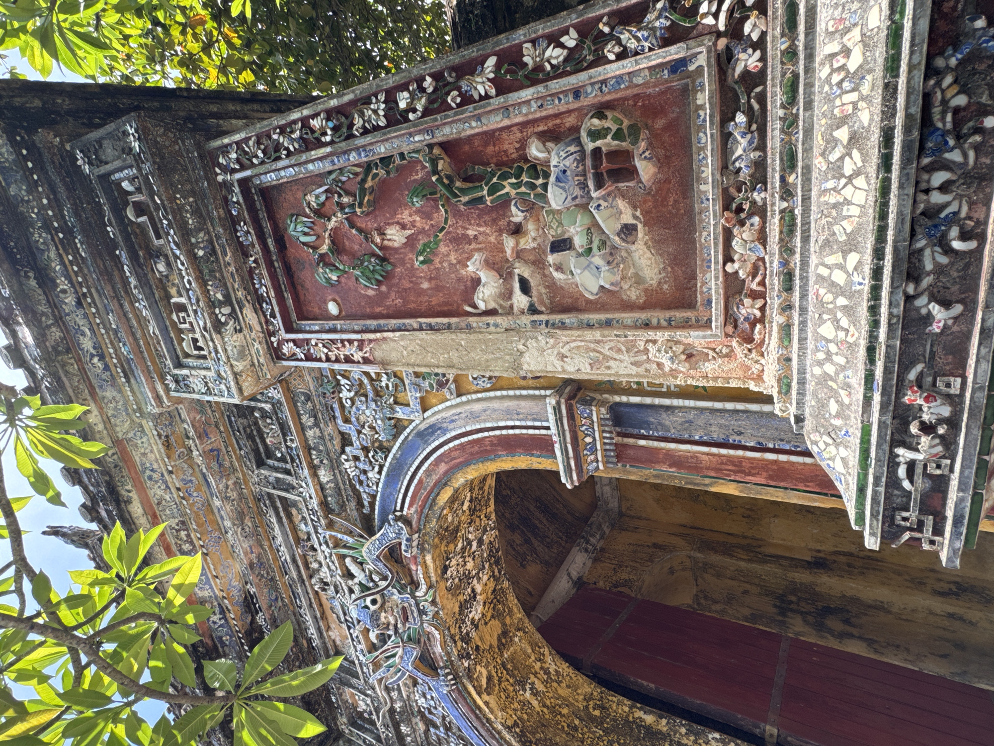 The image shows an ornate architectural detail of a gate, likely part of an ancient structure in Phú Xuân Ward, Vietnam. The gate features intricate mosaic work with vibrant colors and detailed reliefs. A prominent blue dragon sculpture adorns the top of an arched entryway, which is framed by red and yellow painted wooden doors. The surrounding walls are covered in colorful mosaic tiles depicting scenes with animals and flora, suggesting a rich cultural and artistic heritage. Lush green foliage from overhanging trees partially obscures the top of the structure, indicating an outdoor setting under a bright sky. The low-angle perspective emphasizes the grandeur and historical significance of the architecture.
