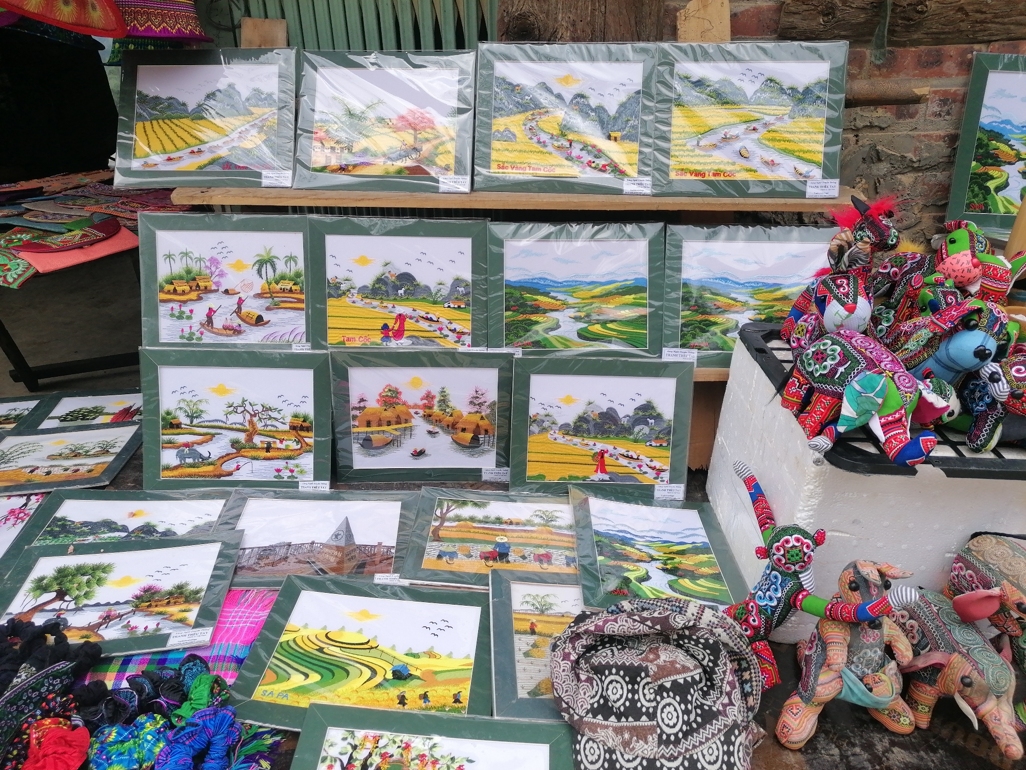 The image captures an array of handcrafted goods for sale, likely in a market setting such as Cat Cat Village, Vietnam. Embroidered pictures framed in green, depicting landscape scenes like rice fields, rivers, and villages, are displayed in rows. Below them, more embroidered art and framed images are spread out. To the right, a pile of stuffed animal-like dolls in vibrant colors and patterns add texture to the display. Scarves or fabric with ethnic patterns can be seen at the bottom. At the bottom left corner, colorful bundles of what might be clothing or other soft goods. A brick wall forms the backdrop on the right, while corrugated metal sheets act as the backdrop on the left side.