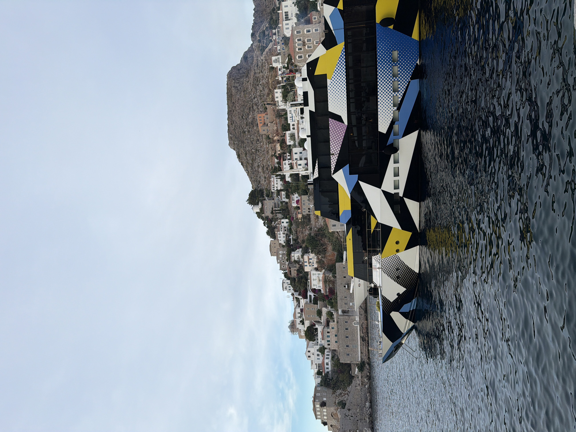 The image depicts a scene in Hydra, Greece. A boat with a distinctive geometric pattern, primarily in black, white, yellow, and blue, is docked along the coast. The boat's reflection is visible in the water. Behind the boat, a hillside is densely populated with white buildings and structures, typical of Greek island architecture. A rocky peak rises above the buildings. The sky is light blue with some cloud cover.