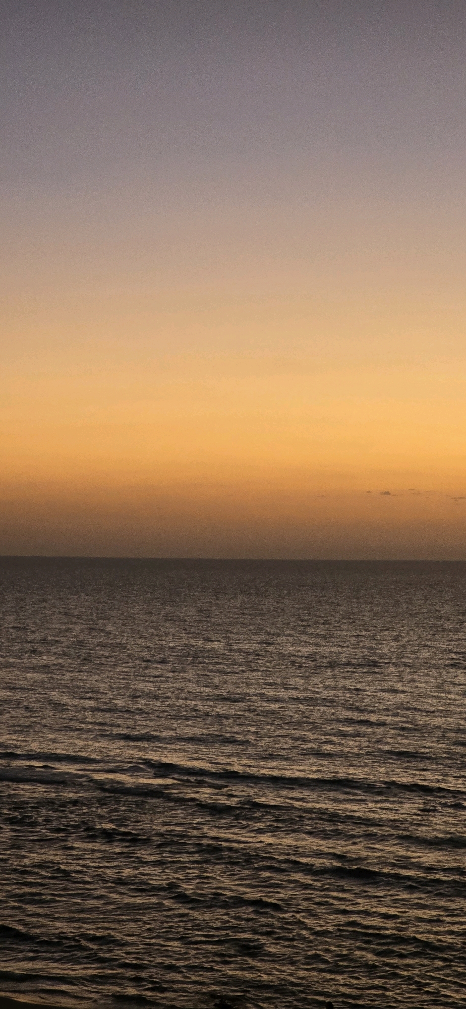 The image depicts a serene seascape at dusk in Bat Yam, Israel. The horizon line is clearly defined, separating the calm, dark, undulating waters of the Mediterranean Sea from the sky above. The sky transitions from a pale purple-gray at the top to a warm, vibrant orange hue near the horizon, indicating the setting sun. The water's surface is textured with small ripples and waves, catching the fading light and reflecting the colors of the sky. There are no people, vessels, or structures visible in the image, presenting a natural and tranquil scene. The overall impression is one of quietude and natural beauty, characteristic of a coastal sunset.