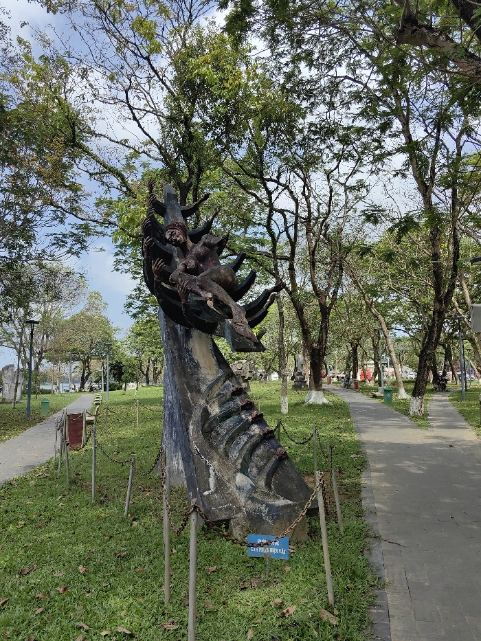 The image displays a public park scene located in Thuận Hóa Ward, Vietnam. A large, weathered sculpture, appearing to be dark grey concrete or stone, occupies the central foreground. This abstract piece features an upward-reaching, jagged form with embedded human figures. One distinct figure, rendered in reddish-brown tones, is visible on the upper section, appearing to be entangled within or emerging from the sculpture's structure. The sculpture's base exhibits horizontal ribbed details and is cordoned off by a low barrier consisting of dark metal posts interconnected by chains. A blue sign with white Vietnamese text, "XIN ĐỪNG LÊN VẬT LẠ" (translating to "Please do not climb on the strange object/thing"), is positioned at the base of the sculpture.
The sculpture is set on a grassy area, sparsely covered with fallen brown leaves. Dark grey paved pedestrian pathways run parallel to the grassy area on both the left and right sides. Along the left pathway, a dark reddish-brown bench-like structure and a green public waste bin are visible, along with a lamppost. The park is densely populated with numerous tall trees featuring green foliage and visible branches against a partly cloudy sky. Several tree trunks bordering the pathways have been whitewashed at their bases. In the background to the left, a body of water, likely a river or lake, is visible, bordered by other less defined sculptures or structures. The background to the right shows more trees, suggesting a broader urban or park environment beyond. No individuals are distinctly visible in the immediate vicinity of the sculpture; distant, indistinct figures may be present further along the pathways.