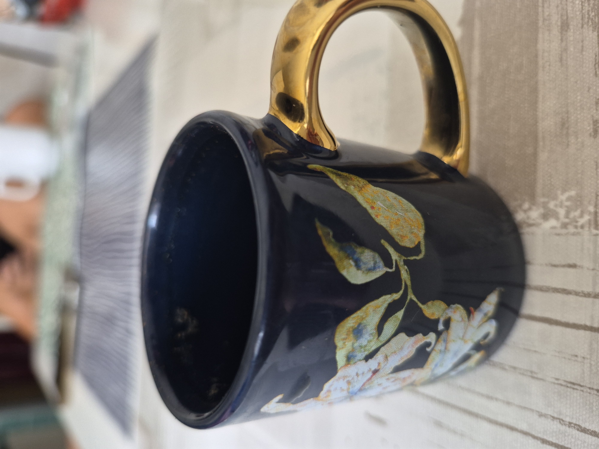A close-up, eye-level shot focuses on a dark blue mug with a gold handle, adorned with a floral pattern featuring light blue and yellow petals. The mug rests on a light-colored, textured surface that appears to be a wooden tabletop with a subtle grain. In the blurred background, hints of a person's arms and torso are visible, along with a white mug and other objects on a surface covered with a patterned mat. The overall lighting suggests an indoor setting, possibly a kitchen or dining area, with soft, ambient light.