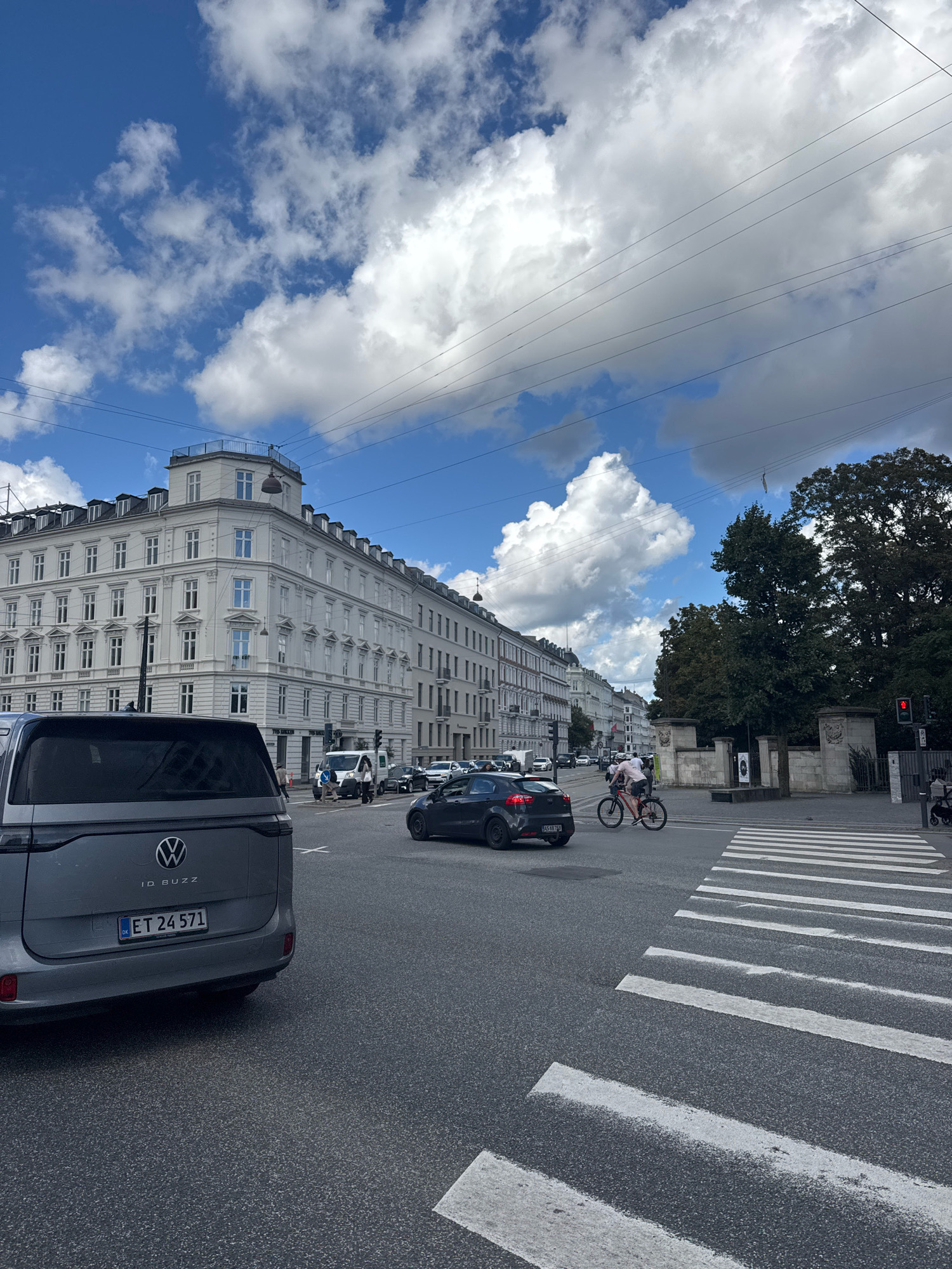 In a city intersection with a clear blue sky and scattered white clouds, a gray Volkswagen ID. Buzz is parked on the left side of the road. The car's rear is visible, showing the license plate "ET 24 571". Across the intersection, a white pedestrian crossing is marked on the asphalt. A person on a bicycle wearing a pink top rides across the crosswalk. In the background, a row of white buildings with multiple windows lines the street, and a large green tree stands to the right of the intersection. Traffic lights are visible, with one red light illuminated.