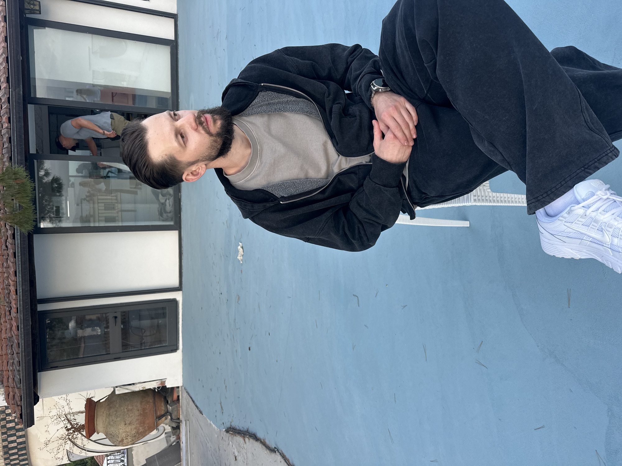 A medium, long shot presents a male in the foreground seated on a white chair on a blue surface, ostensibly a patio. The male wears dark grey sweatpants, a dark grey zip-up hoodie, and white sneakers. He has a beard. His hands are clasped.
In the background, through a glass-paneled door, another male figure is visible. He appears to be bending over or cleaning something, wearing a grey shirt and olive pants.
Architectural elements suggest an Italian villa. A clay pot is visible, and a tile roof appears in the background. Given the architectural details, this is likely Taormina, Italy. The weather is daytime.
