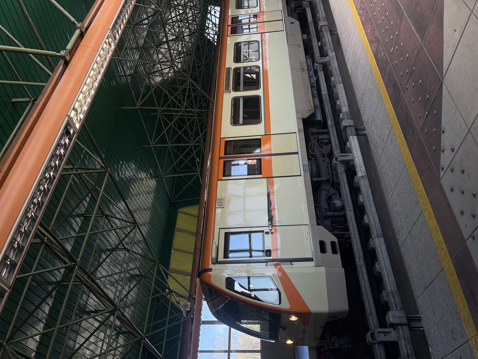 The image captures a train carriage at the Serdika metro station in Sofia, Bulgaria. The train is positioned at a platform, alongside a tiled floor with tactile paving. The station has an overhead green structural framework and integrated lighting. The carriage itself is predominantly cream in color, accented with orange stripes and multiple windows. The exterior environment is faintly visible through the front window of the train.