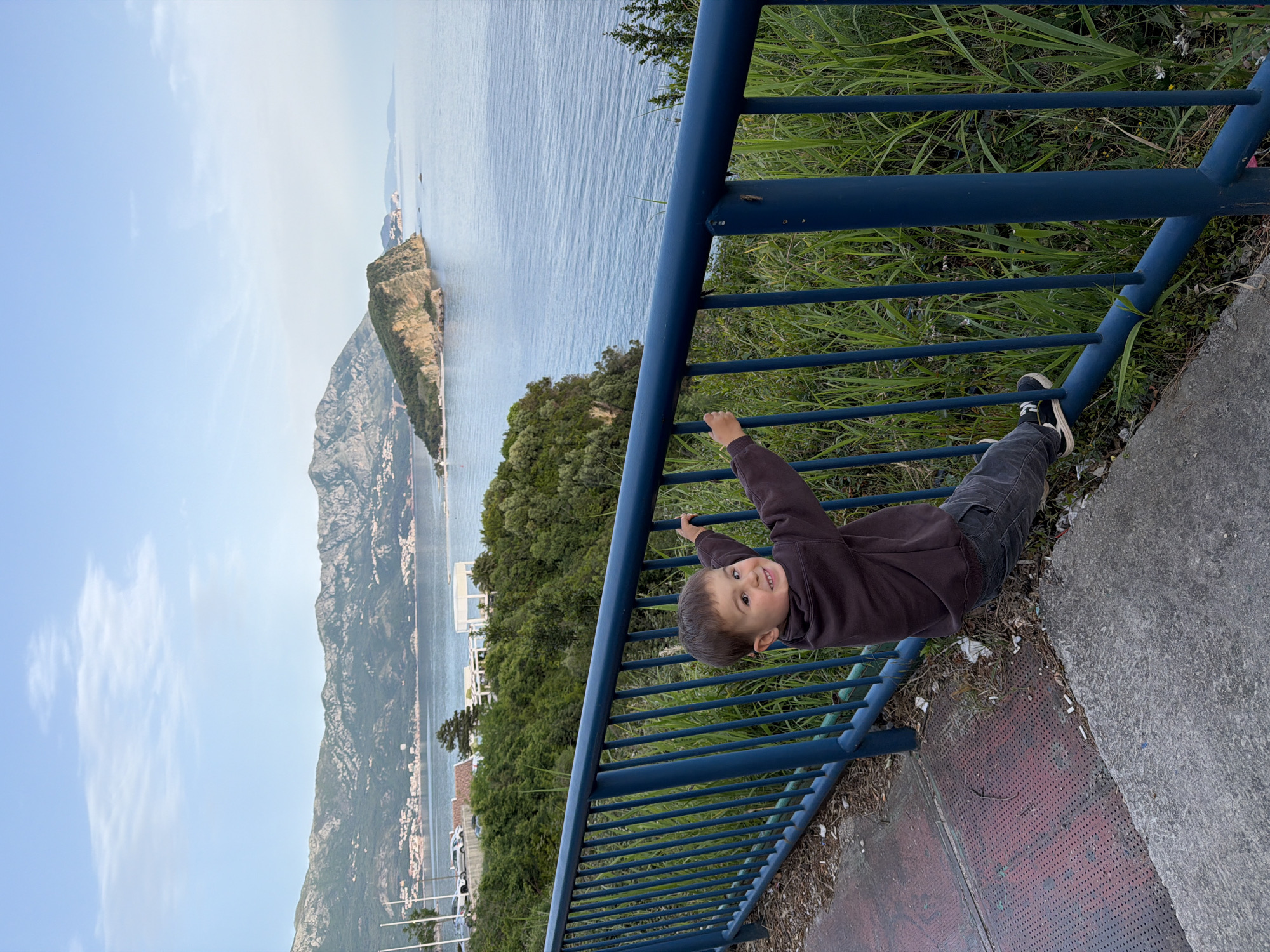 A child is hanging upside down from a blue metal railing. The child is wearing a brown sweatshirt and dark pants with black and white sneakers. The child is smiling and looking toward the viewer. The railing is located along a pathway next to a grassy area. Beyond the railing and grass, there is a view of the Adriatic Sea, a mountainous coastline, and a partial view of the city of Budva, Montenegro. The sky is pale blue with light clouds. The photograph is rotated clockwise approximately 90 degrees.