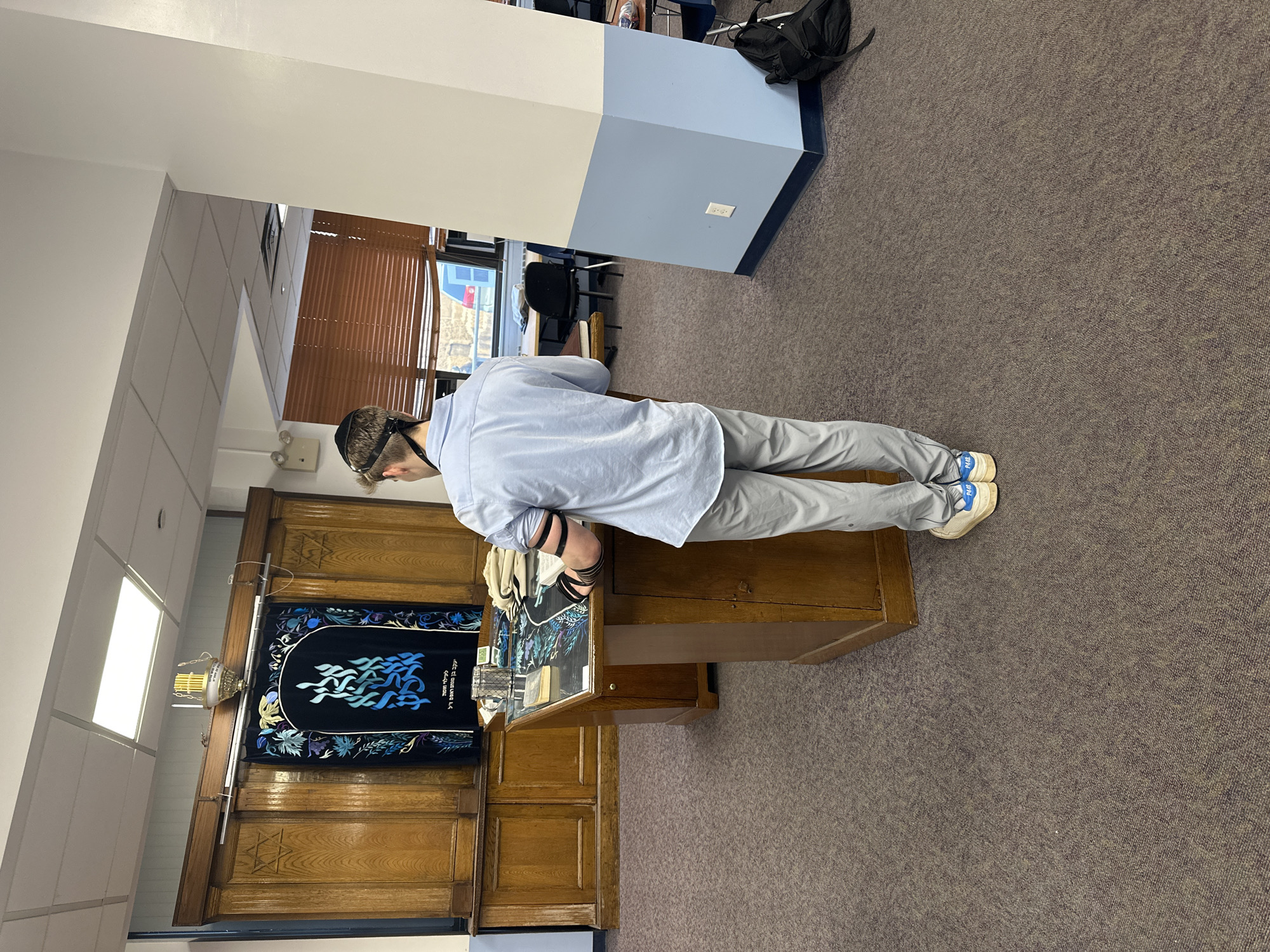 An interior photograph depicts a young male, viewed from behind, engaged in morning prayers within what appears to be a synagogue or study hall. The male, wearing a light blue button-up shirt, grey pants, and light-colored sneakers, has tefillin (phylacteries) wrapped around his left arm and on his head, beneath which a kippah is visible. He stands before a wooden lectern with a slanted top, looking down at an open book. An ornate metal cup and a light-colored cloth bundle rest on the lectern's surface, which features a decorative overlay.
In the background, a prominent wooden Aron Kodesh (Holy Ark) is visible, featuring ornate paneling with two carved Stars of David. A dark blue or black velvet Parochet (Ark curtain), embroidered with light blue and white stylized flames and Hebrew text including "אש תמיד" (Eternal Flame) and memorial dedications, hangs over the ark's opening. Above the ark, a brass Ner Tamid (Eternal Light) is suspended.
The room features a patterned brown carpet and a white drop ceiling with square fluorescent light fixtures. To the right, a two-toned pillar (white top, light blue bottom with a dark blue base trim) stands in the foreground. Beyond the pillar, tables and chairs are visible through a window covered by brown Venetian blinds, offering a partial view of an urban exterior with a blue vehicle. A black backpack rests on the carpet next to the pillar. While the setting is characteristic of Jewish religious institutions prevalent in New York City, a specific location within the city cannot be identified from the visual evidence.