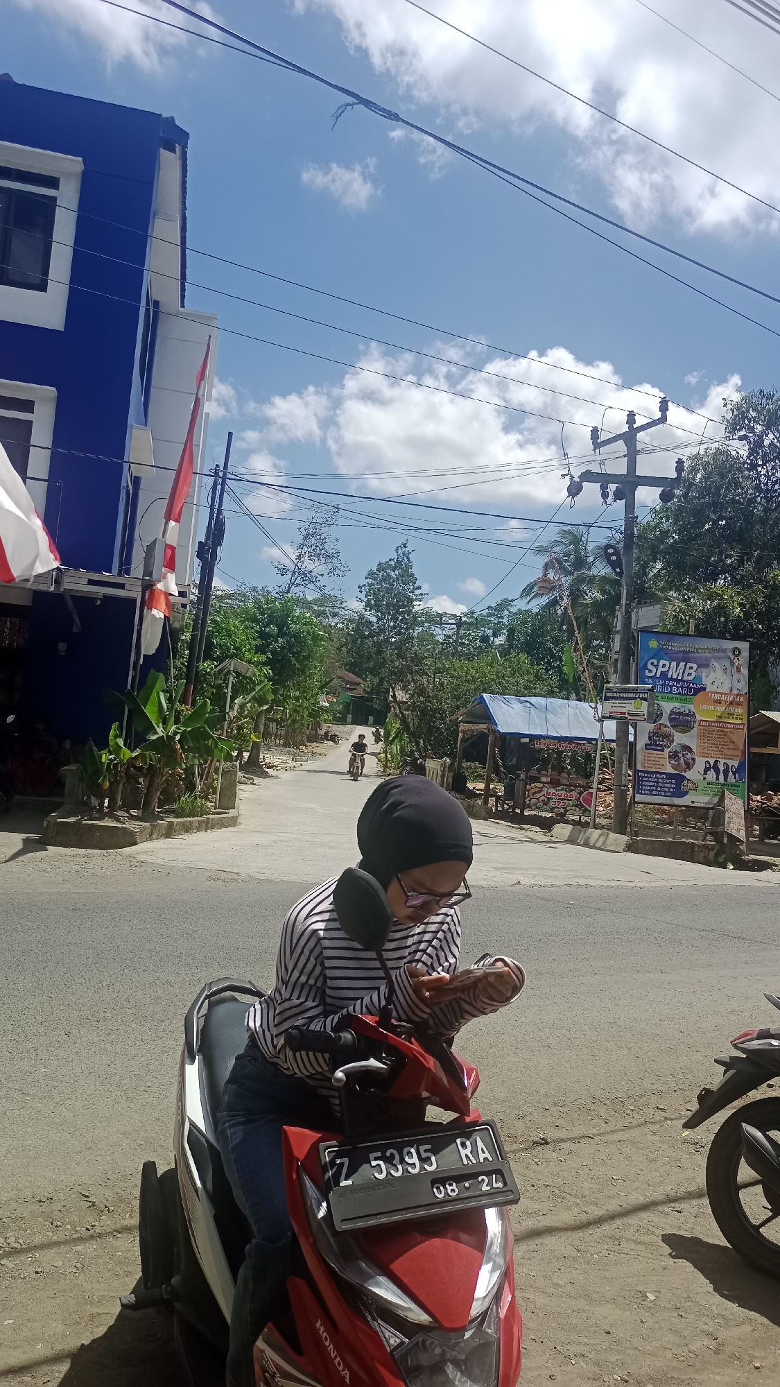 A young woman wearing a black hijab and a striped shirt is sitting on a red scooter on a paved road in Cibeber, Indonesia. She is looking down at her phone, which she is holding in her hands. The scooter has a license plate that reads "Z 5395 RA". In the background, there is a blue building with white trim, a red and white flag, and several trees. A banner for "SPMB Sistem Penerimaan Mahasiswa Baru" is visible on a utility pole. The sky is blue with some white clouds. There is another scooter parked to the right of the frame. The scene appears to be a street in a somewhat rural or suburban area.