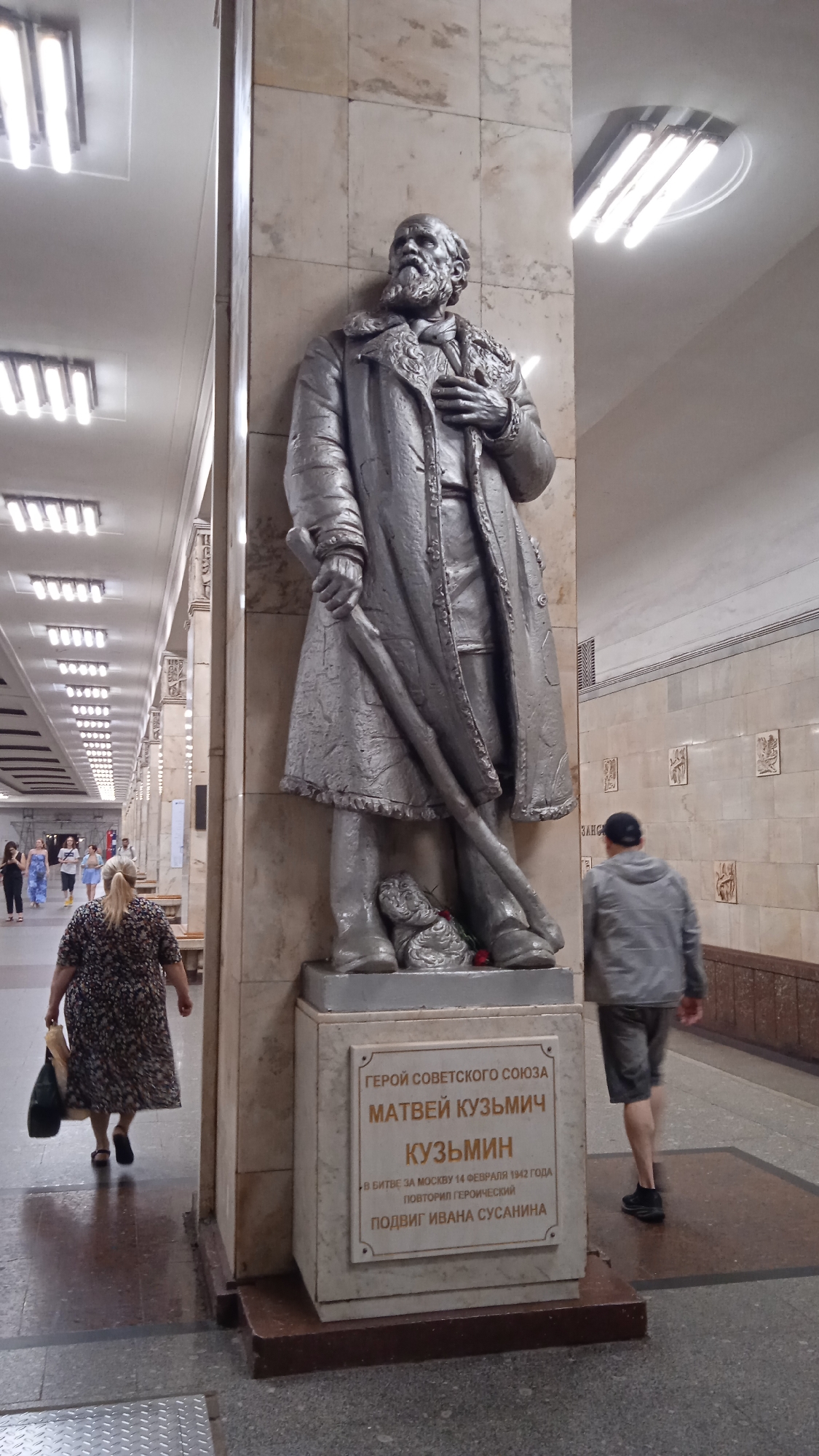 A statue of Matvey Kuzmich Kuzmin, a Hero of the Soviet Union, stands in a Moscow Metro station. The statue depicts Kuzmin in a long coat and hat, holding a rifle. A plaque at the base of the statue reads: "Hero of the Soviet Union Matvey Kuzmich Kuzmin. In the Battle for Moscow on February 14, 1942, he repeated Ivan Susanin's heroic deed." A woman with blonde hair walking away from the camera is in the foreground, and a man in a grey jacket and shorts is walking past the statue in the background. The station is decorated with marble walls and fluorescent lighting.