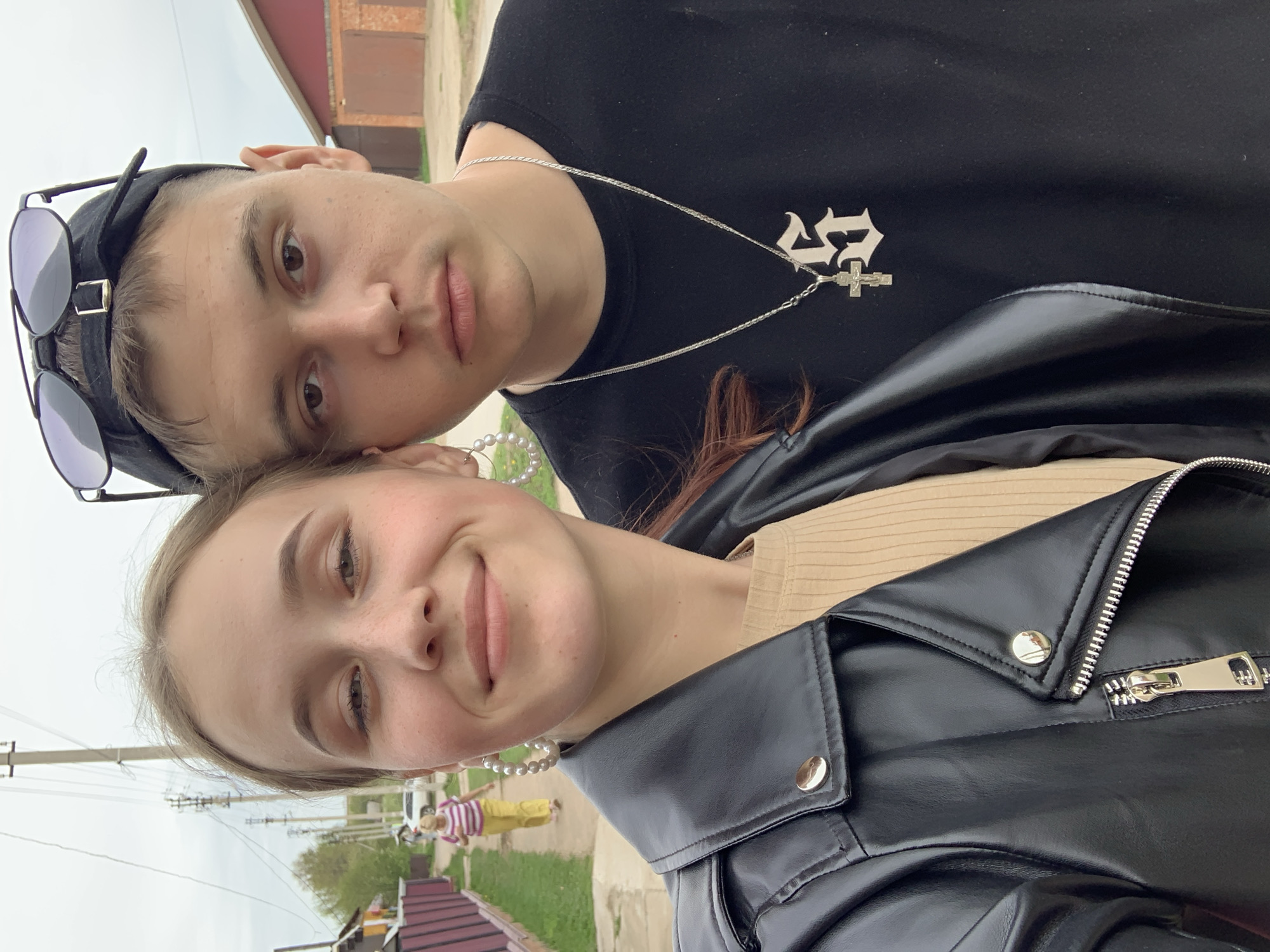 A selfie photograph depicts two individuals, likely a male and female, outdoors. The female, with light skin and brown hair, is positioned in the foreground, wearing pearl earrings and a black leather jacket over a tan shirt. She is smiling slightly at the camera. Behind her is a male with light skin and short, dark hair, wearing a black T-shirt with a silver chain and cross pendant, along with a black cap with sunglasses perched on it. He has a more neutral expression. The background includes a sidewalk and a street scene with a wooden house, a utility pole with wires, and trees. There is a blurred figure in the distance wearing a striped shirt and yellow pants. The photo is taken during the daytime with overcast lighting. Given the objects and landscape, it is most likely a rural area of the city of Фоки, Russia.