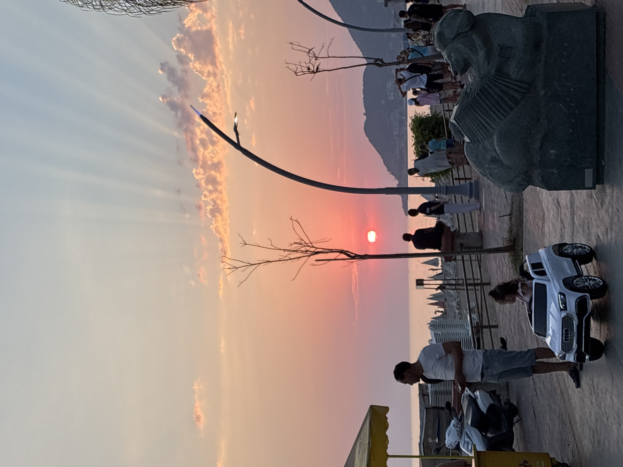 At sunset in Alanya, Turkey, the sky is painted with hues of orange and pink, with crepuscular rays emanating from the setting sun. A group of people are gathered along a promenade overlooking the sea. In the foreground, a man stands next to a white toy car with a young girl inside, while a scooter and a yellow umbrella are visible to the left. Further along the promenade, various individuals are walking and socializing. A distinctive sculpture of a dog playing an accordion sits on a pedestal. The background shows the coastline and hills of Alanya.