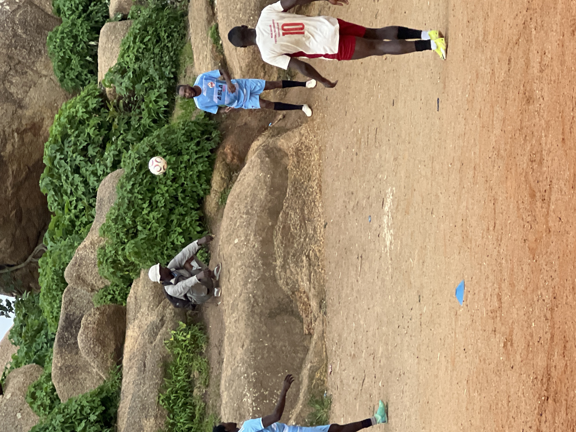 The image shows a group of young men playing soccer in a rocky, outdoor setting. The scene appears to be in Jos, Nigeria. Three individuals are visible in active or semi-active poses. One person, wearing a white jersey with the number "10" and red shorts, is in mid-stride, suggesting they are running. Another person, in a light blue soccer jersey with "ASLA" written on it and blue shorts, is standing, looking towards the ball. A third person, dressed in a grey jacket, white hat, and dark pants, is sitting on a rock, seemingly observing or resting. A soccer ball is visible in the air, mid-play. The ground is a mixture of dirt and scattered small stones, with large, weathered rocks and lush green vegetation forming a natural backdrop. The lighting suggests it is daytime, possibly late afternoon given the warmth of the light. The overall atmosphere is casual and recreational, indicating a friendly soccer match or practice.