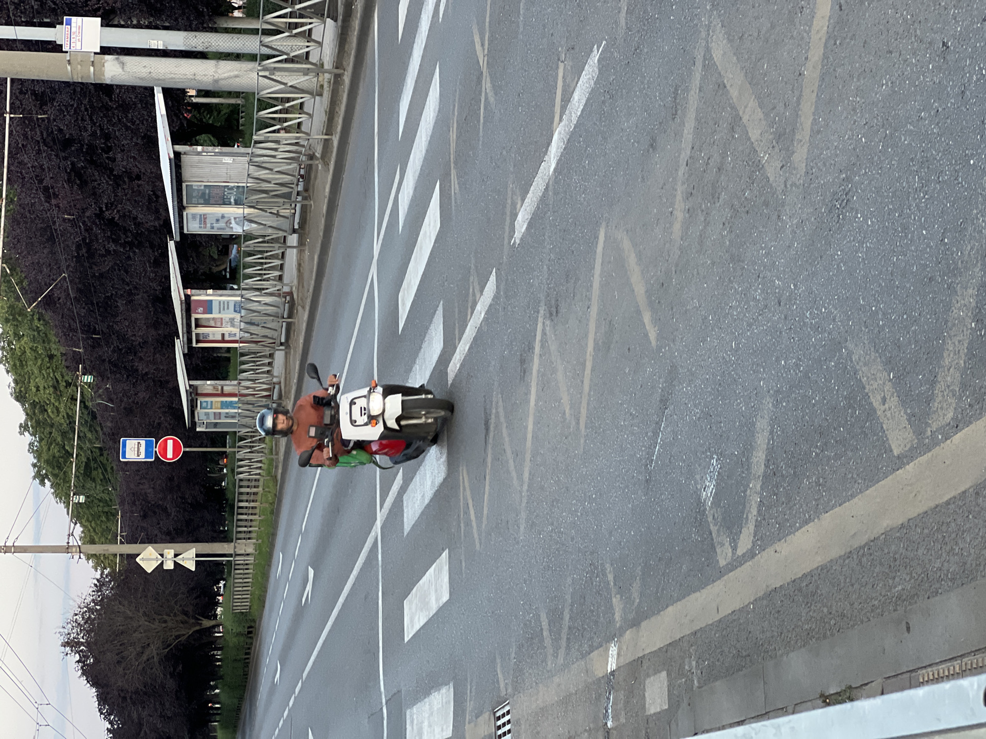 A person wearing a red shirt and a dark helmet rides a white and red scooter through a pedestrian crossing. The scooter has a green bag attached to the back. The scene takes place on a multi-lane road in Krasnodar, Russia, indicated by traffic signs including a bus stop sign and a red circular "no entry" sign. The background features a row of dark trees and bus shelters. The road surface is asphalt, with white lane markings and yellow edge lines. Overhead power lines are visible.