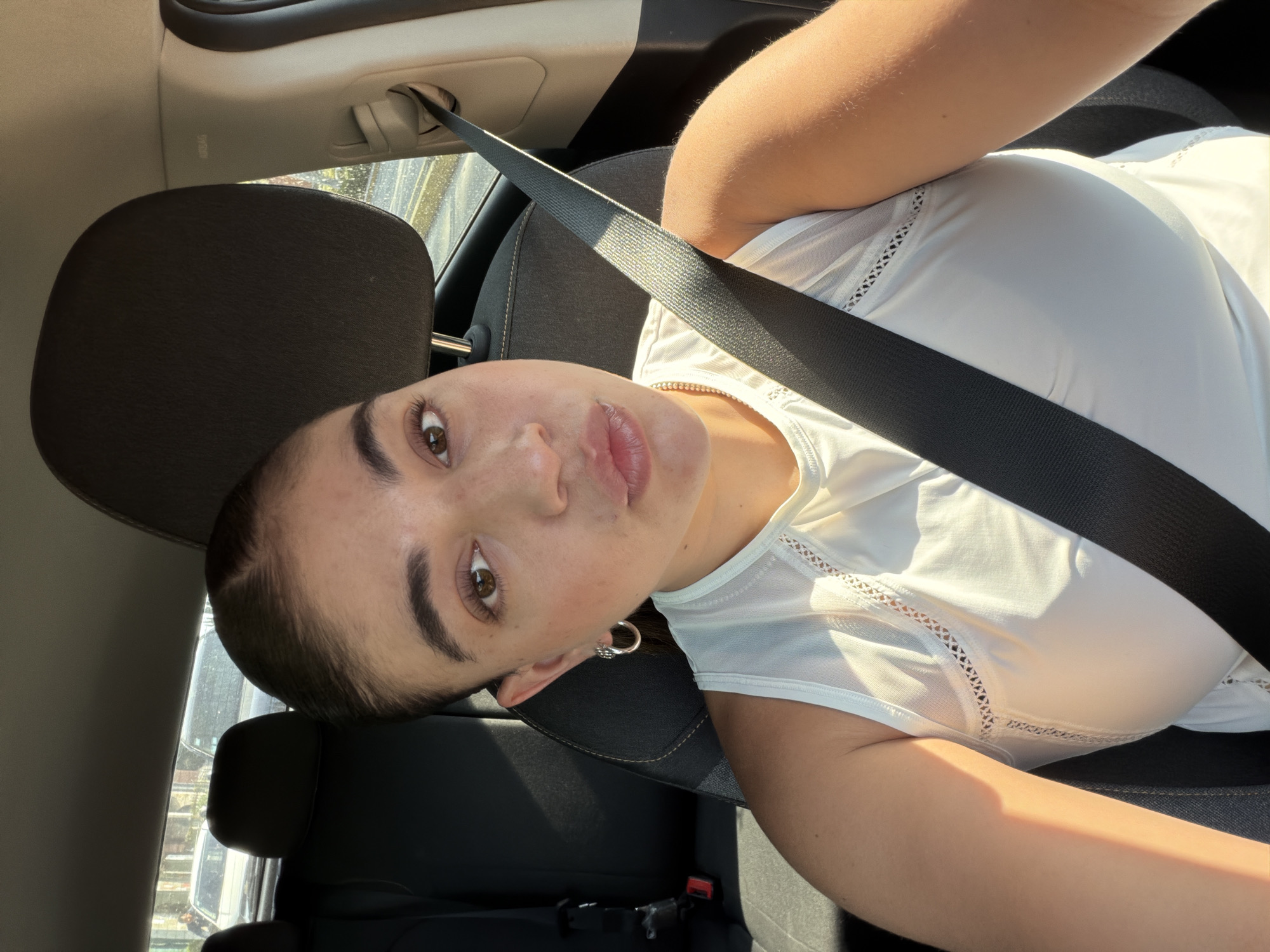 The image shows a woman wearing a white shirt and a seatbelt, inside a car. The car's interior is dark. She is looking at the camera. The image appears to have been taken from an unusual angle, with the camera tilted. The location is not discernible from the image. There is no evidence the photo was taken in Puebla, Mexico.