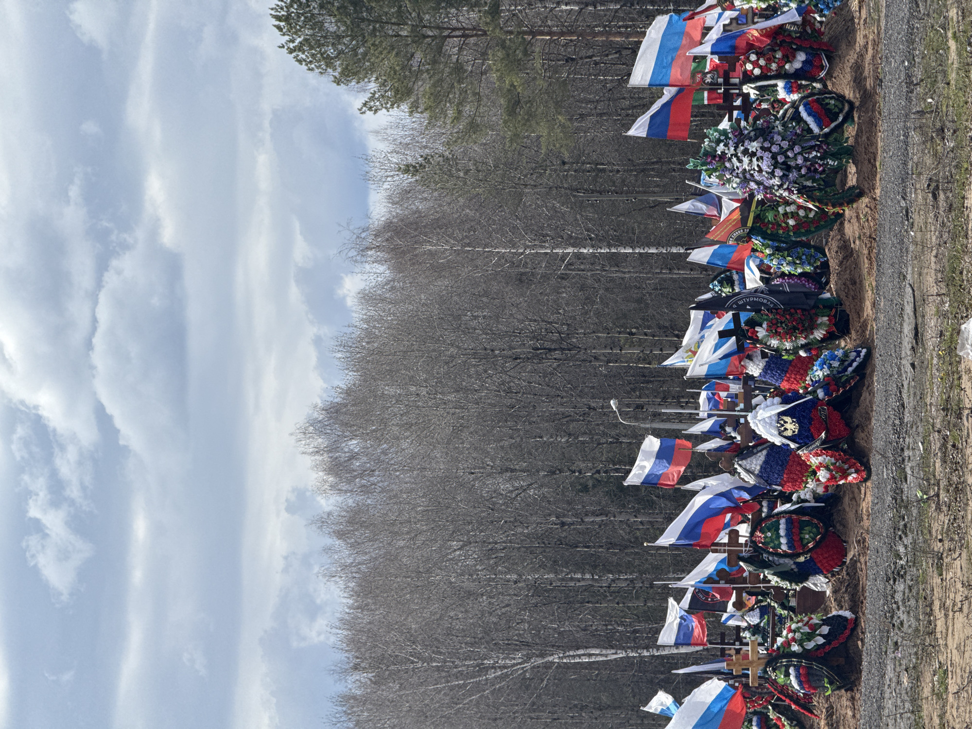 The image displays a somber outdoor scene, likely a military cemetery or a memorial site in Perm, Russia. The photograph appears to have been taken with a vertical orientation but is presented here rotated horizontally.
In the foreground, a long, neat row of graves or memorial markers stretches across the frame. Each marker is adorned with multiple flags and floral wreaths. Numerous Russian national flags, characterized by white, blue, and red horizontal stripes, are prominently planted on poles. Among these, other flags and banners are visible, including one with black, blue, and red stripes, and another resembling the St. Andrew's flag (Russian Navy ensign).
A multitude of wreaths, both artificial and possibly natural, are placed on the ground, many incorporating the colors of the Russian flag (red, white, and blue), alongside green, black, and purple elements. Interspersed with the flags and wreaths are simple wooden crosses, marking individual burial sites.
Several pieces of text are visible on banners and wreaths. On a black banner, the word "ШТУРМОВАЯ" (SHTURMOVAIA), meaning "Assault" or "Storm," is legible. Another banner partially reads "... ОТЕЧЕСТВУ" (... OTECHESTVU), meaning "... to the Fatherland." Most notably, on a small black flag or banner, the abbreviation "P.M.C." (Private Military Company) followed by "ГРУППА 39" (GRUPPA 39 - Group 39) is clearly visible, indicating a connection to private military formations.
The ground immediately in front of the markers is bare earth or gravel. In the background, a dense forest of mostly bare deciduous trees, with some evergreen pines, stands under a light blue sky dotted with scattered white clouds. The diffuse lighting suggests a daytime setting, likely early spring or late autumn given the lack of foliage on many trees. No people are present in the image, contributing to the quiet and solemn atmosphere of remembrance. A light pole is faintly visible on the far right.