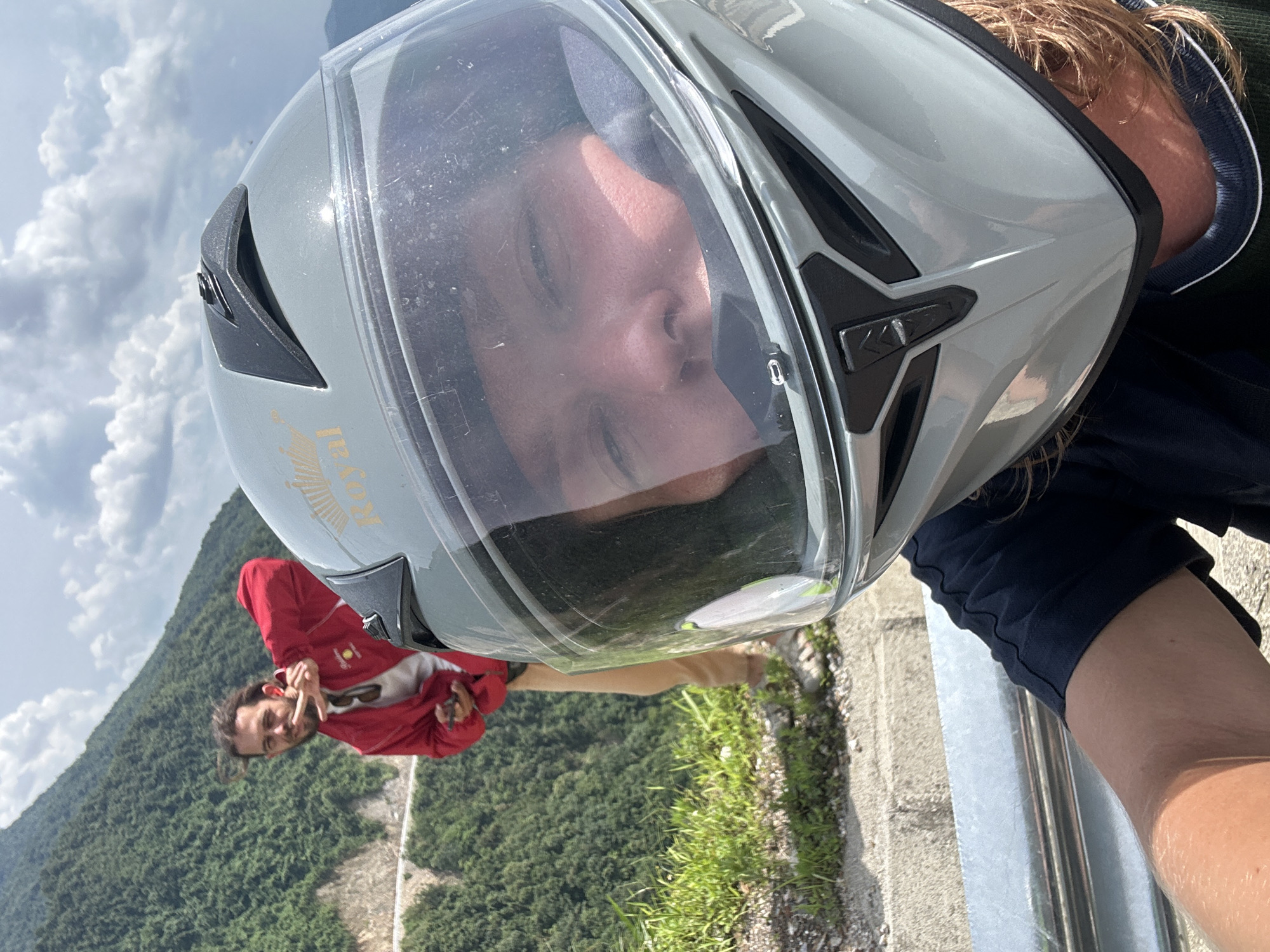 This rotated portrait image captures a close-up selfie or POV shot, set against a mountainous landscape in Muong Than, Vietnam.
In the foreground, the primary subject is seen from a first-person perspective, wearing a light grey full-face motorcycle helmet with a clear visor. The visor reflects the person's face, showing a neutral or slightly smiling expression. Wisps of light brown or blond hair are visible peeking out from under the helmet, which also has a faint gold "Royal" logo with a crown on its side. The person is wearing a dark blue or black top, and an arm, covered in the same fabric, is visible along a metallic barrier in the lower right.
In the mid-ground, a second person, a male with dark hair and a beard, is visible. He is wearing a red long-sleeved jacket, possibly over a light-colored undershirt, and light-colored trousers. He is smiling and making a hand gesture, possibly a peace sign.
The background reveals a vast, lush green mountainous region, densely covered with trees, typical of the Vietnamese landscape. A road or path can be seen winding through the lower part of the mountains in the distance. The sky above is bright with scattered white cumulus clouds. The scene appears to be taking place on a bright, possibly partly cloudy, day, suggesting a stop during a journey, likely a motorcycle ride, to enjoy the scenic view.